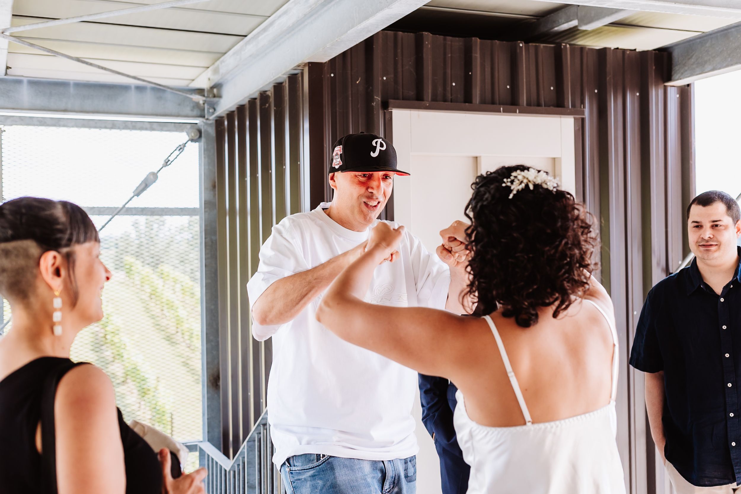 Man in white shirt and cap kisses a bride's hand at an indoor wedding celebration.