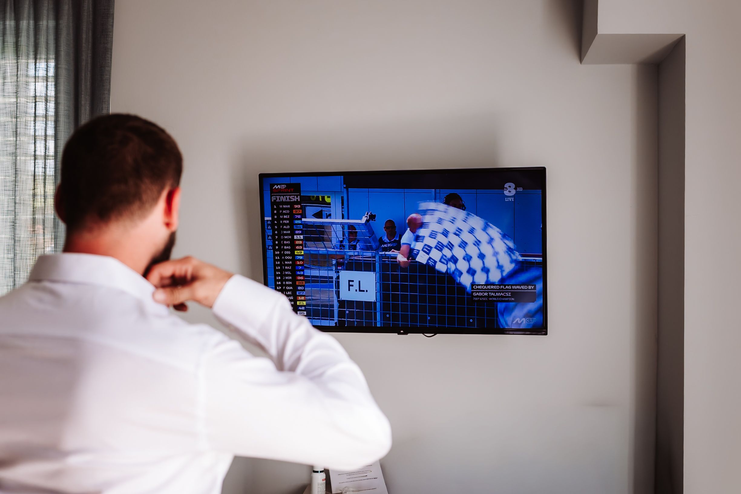Man in white shirt watching a flat screen TV mounted on wall displaying a racing video game with checkered flag.