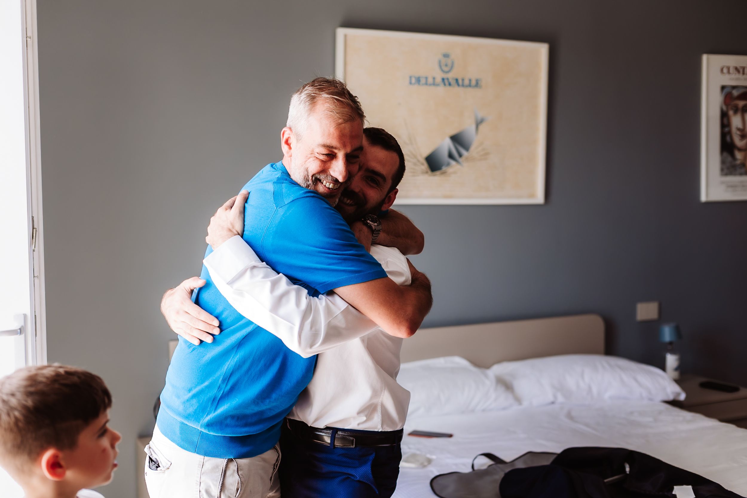 Two men sharing a warm hug in a hotel room, with a third person visible in the foreground.