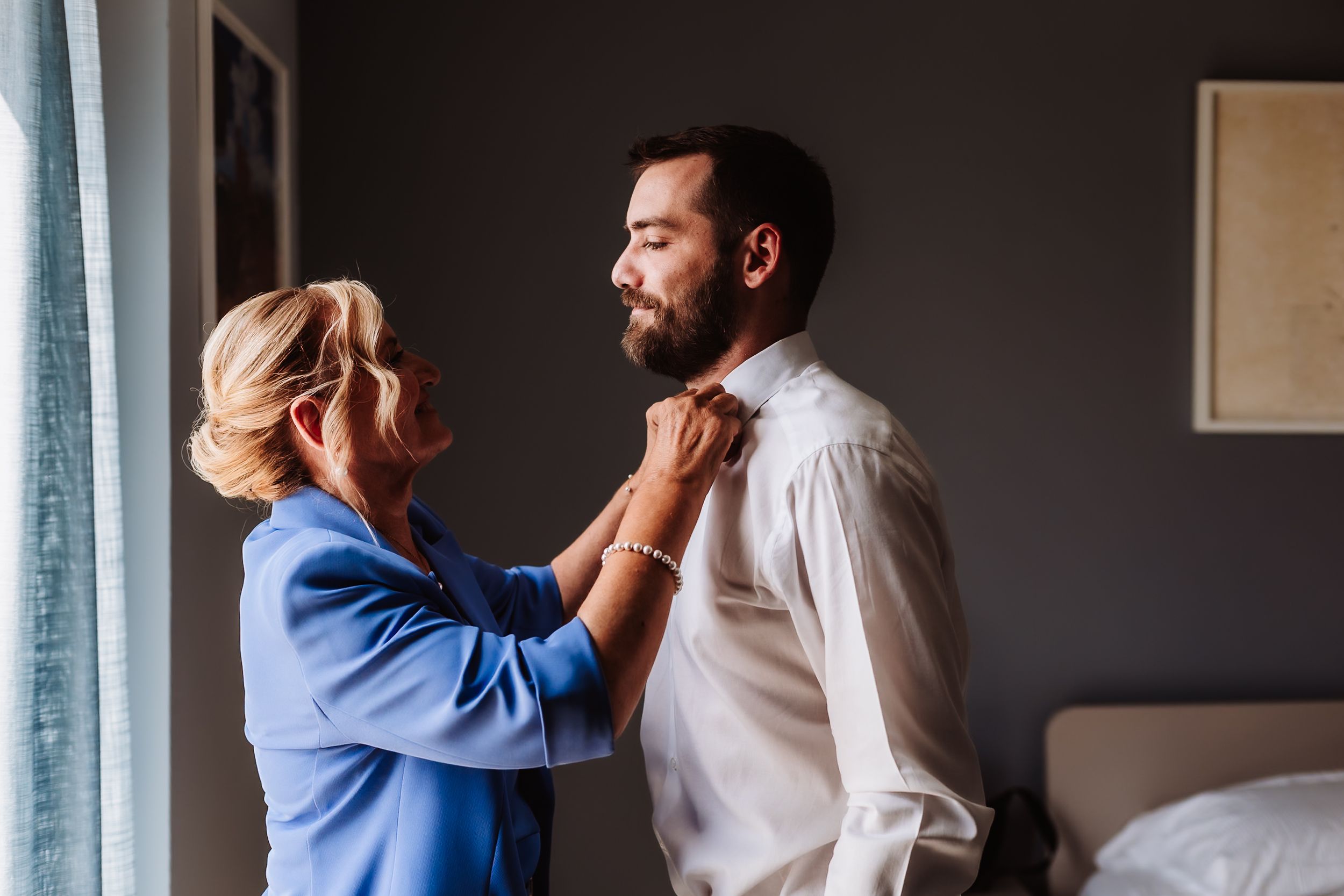 A woman in blue adjusts a bearded man's shirt collar in a softly lit room, likely preparing for a formal event.