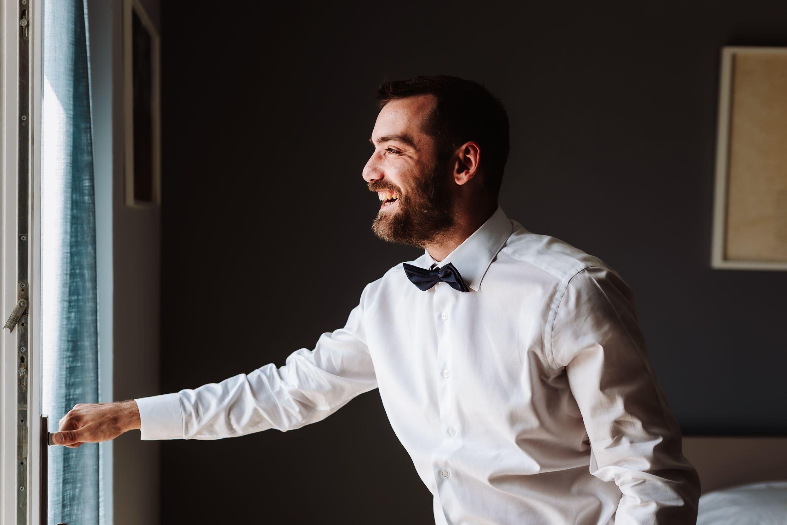 Smiling groom in white shirt and navy bow tie gazes out a window on his wedding day.