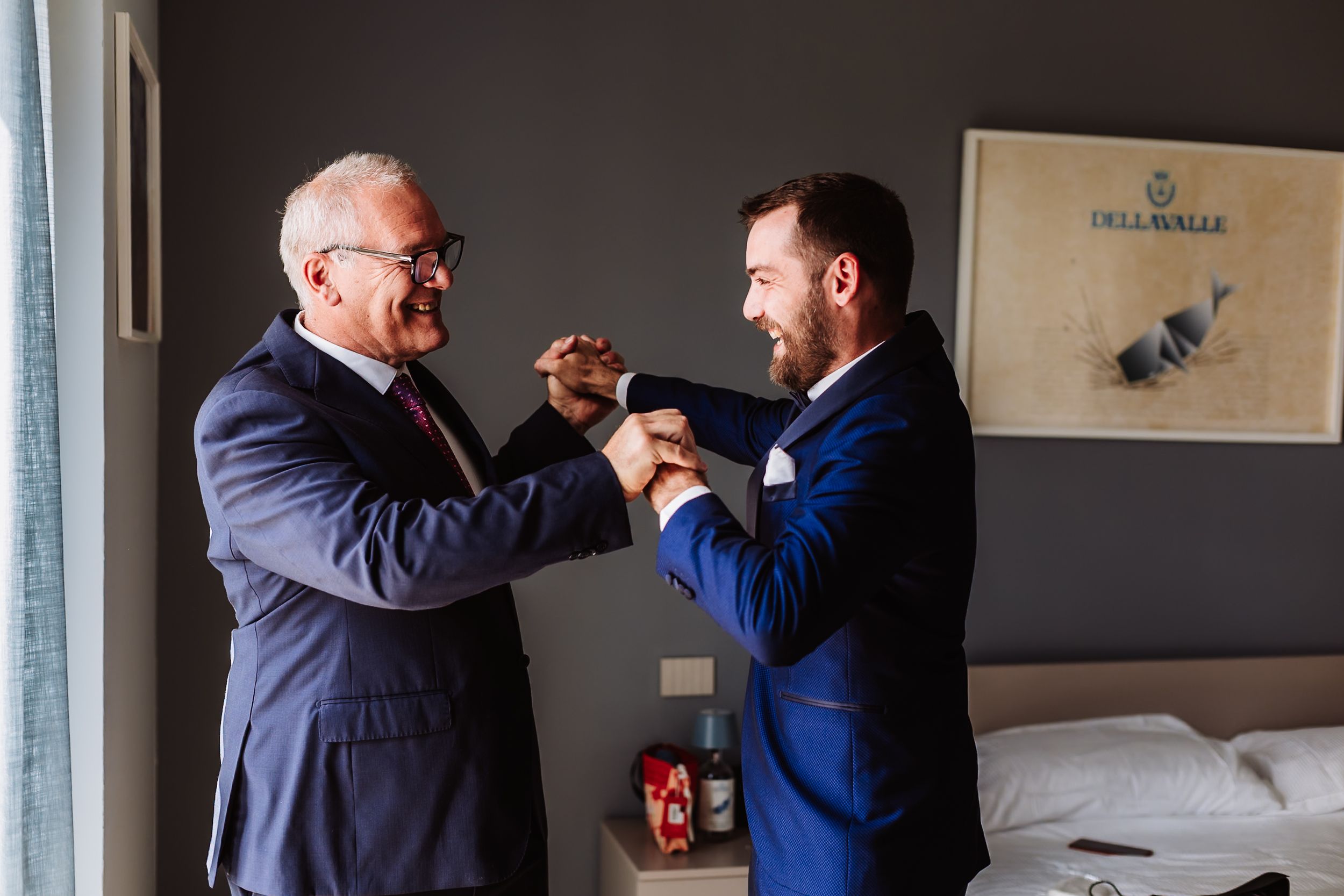 Two men in navy suits sharing a joyful fist bump moment while getting ready for a wedding celebration.