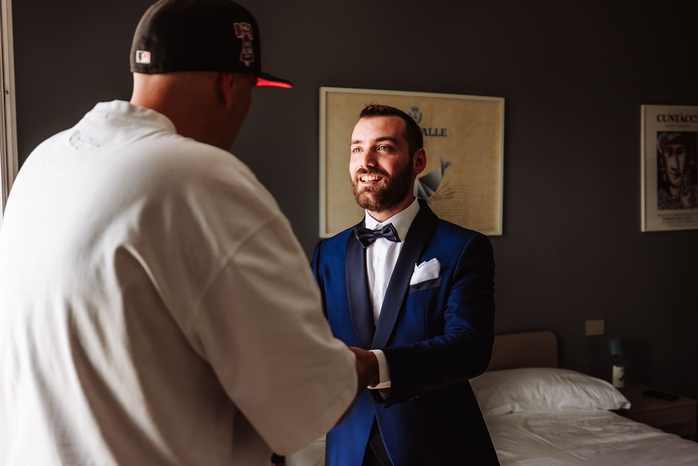 Groom in blue tuxedo smiling and shaking hands with groomsman in hotel room before wedding ceremony.