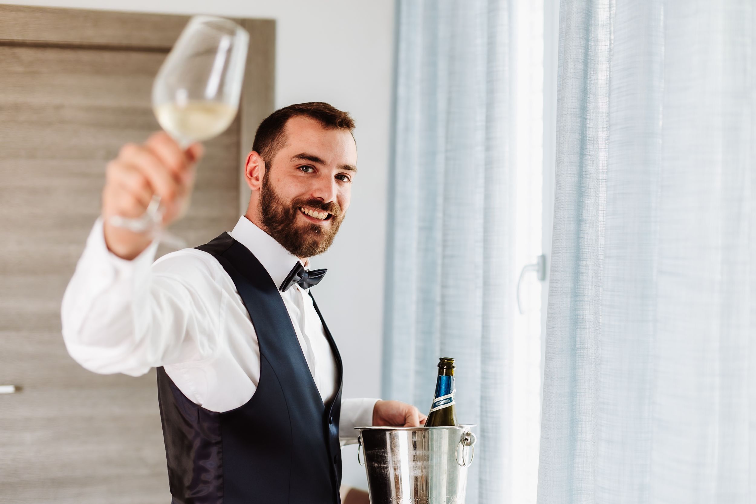 Smiling bearded man in vest and bow tie raising a champagne glass near a bottle in an ice bucket.