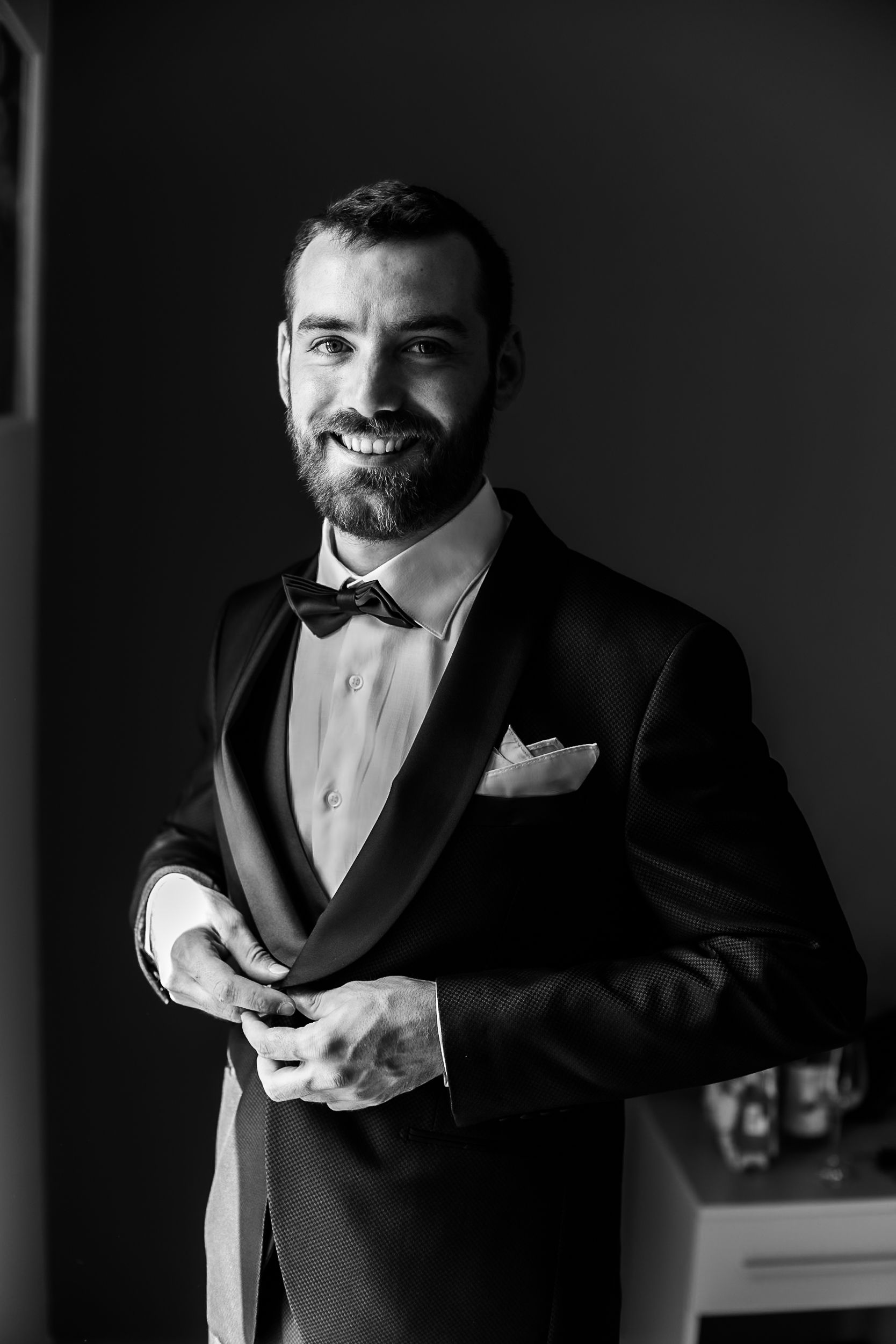 Dapper bearded groom in black tuxedo with bow tie and pocket square smiling in black and white photo.