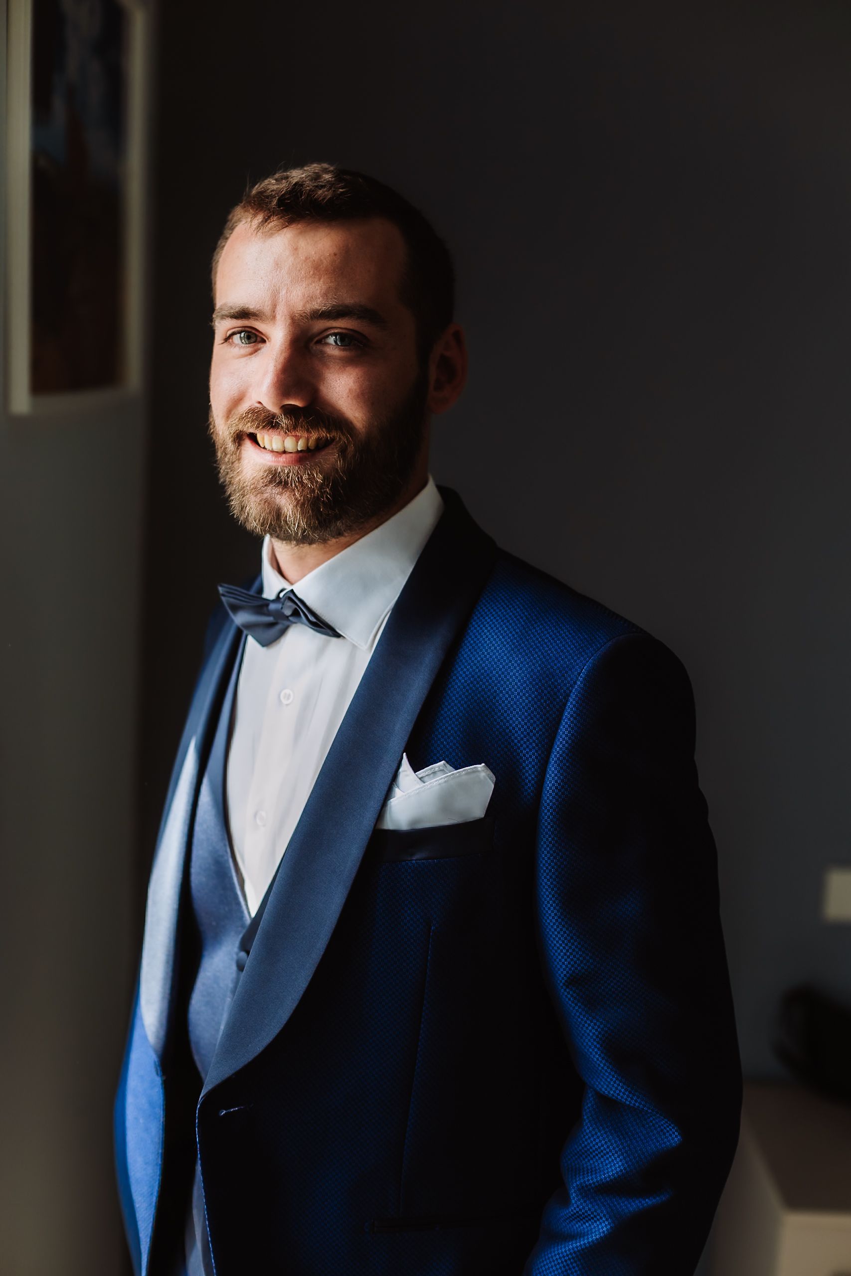 Smiling groom in a navy blue tuxedo with bow tie and white pocket square posing for a wedding portrait.