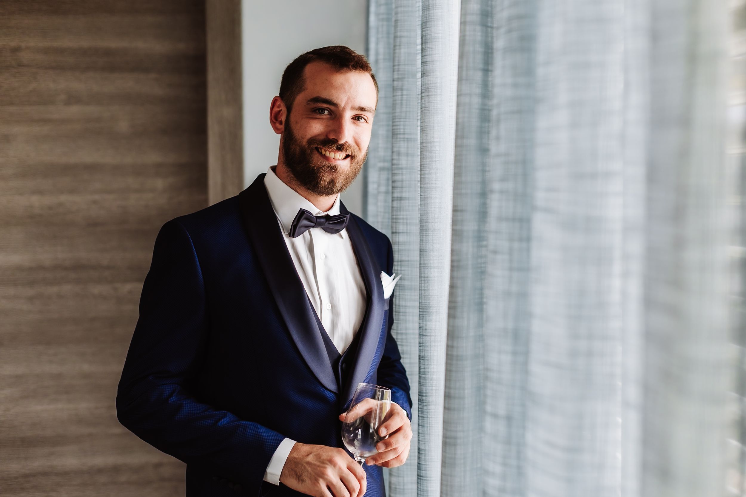A smiling groom in a navy tuxedo with bow tie holds a glass near a window on his wedding day.