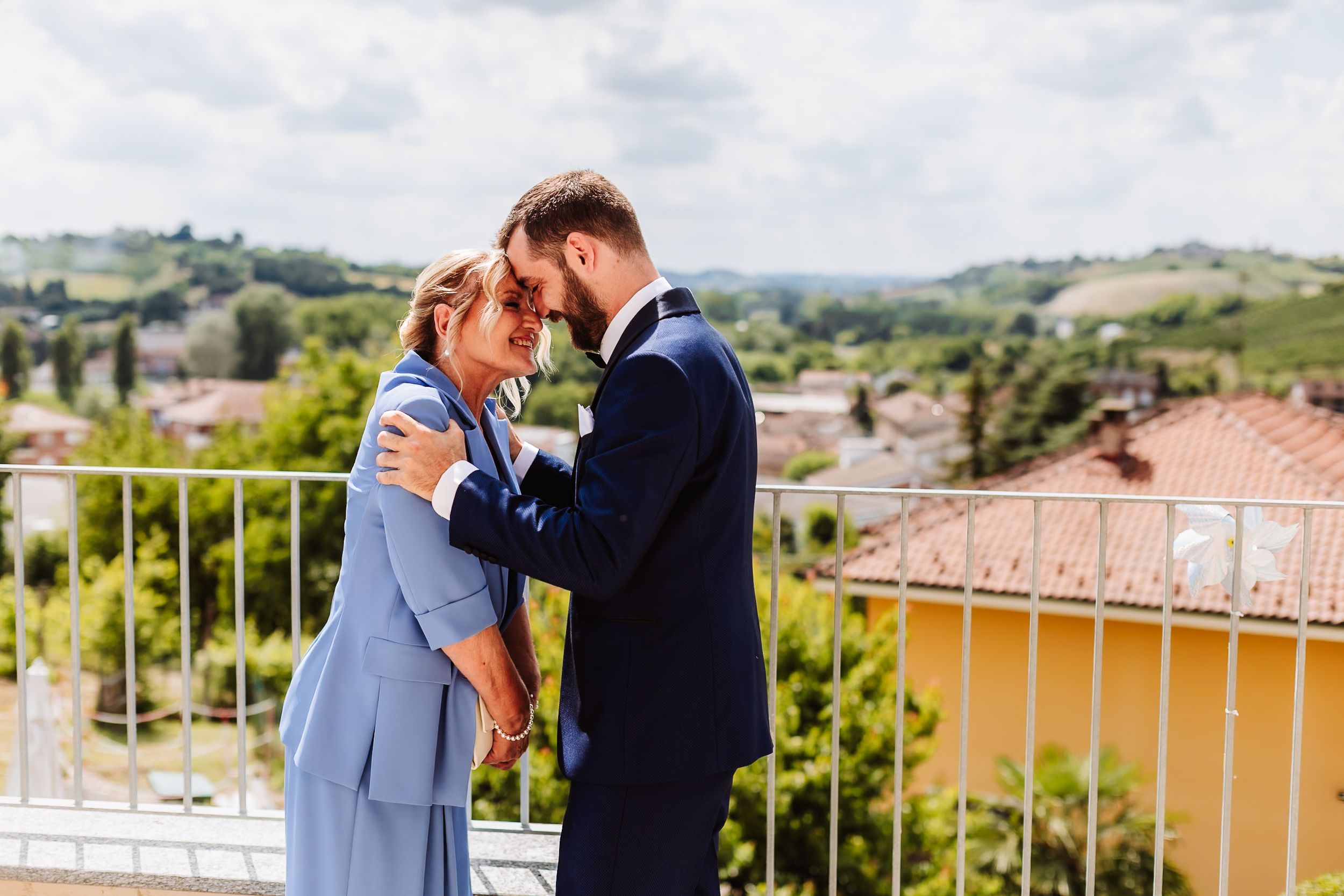 Couple in elegant attire embrace on a balcony with scenic Italian countryside and terracotta rooftops behind them.