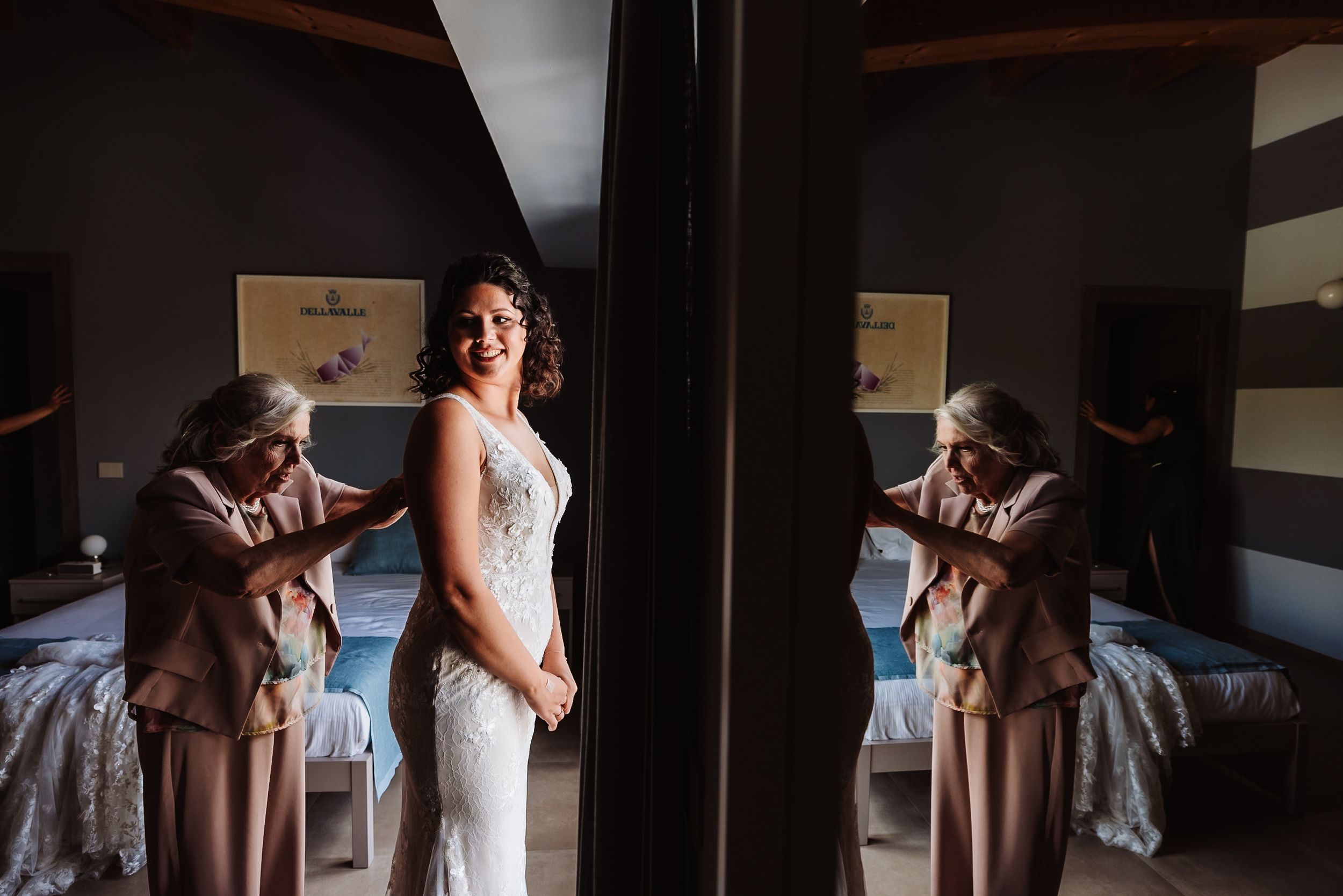 Bride in white lace gown stands by mirror as older woman helps with dress in elegant bedroom setting.