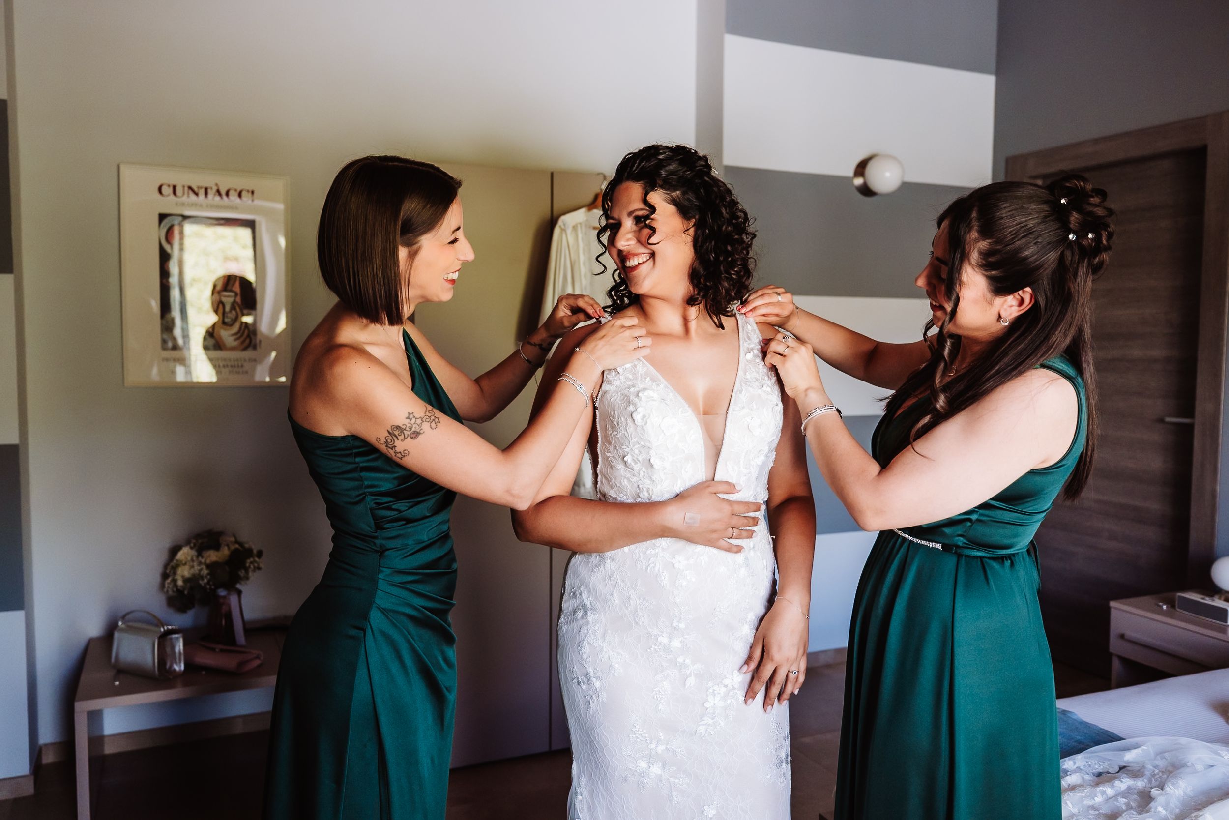 Bride in white lace gown assisted by two bridesmaids in emerald green dresses during wedding preparation indoors.