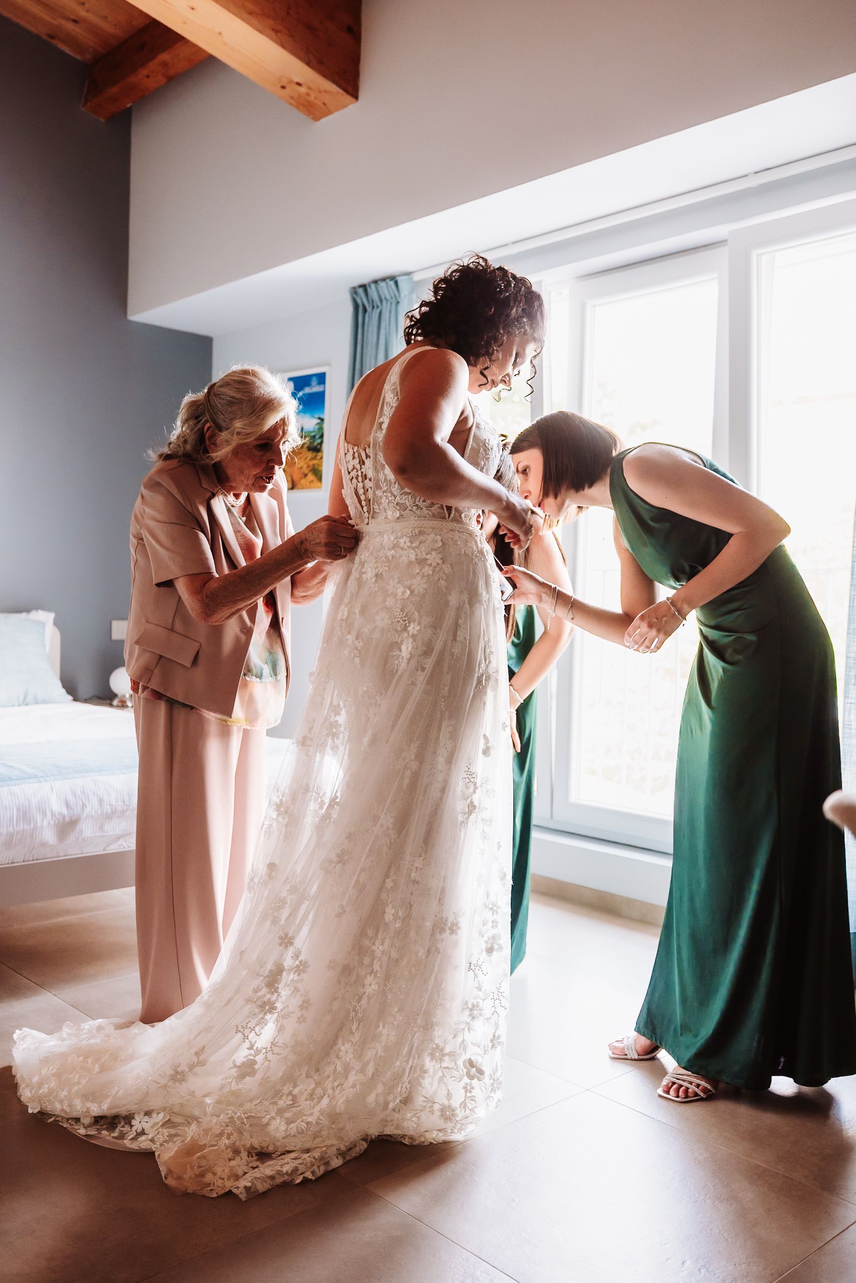 Bride in lace wedding gown being helped by mother and bridesmaid in green dress during wedding preparation indoors.