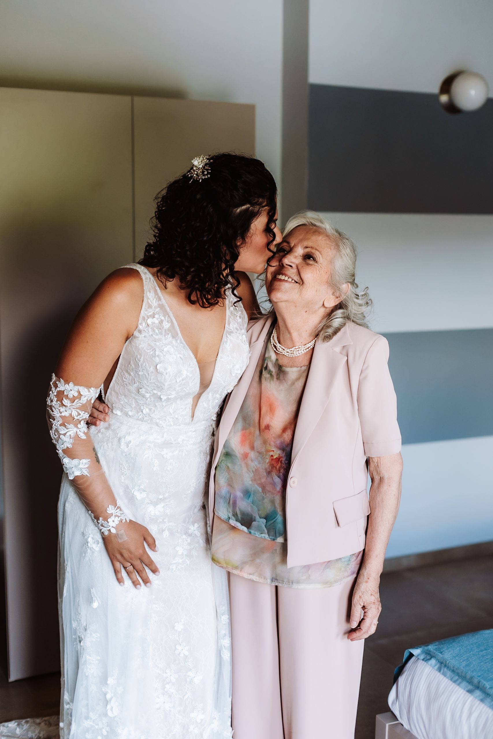Bride in white lace gown kissing elderly woman in pink suit on the cheek, sharing a joyful wedding day moment.