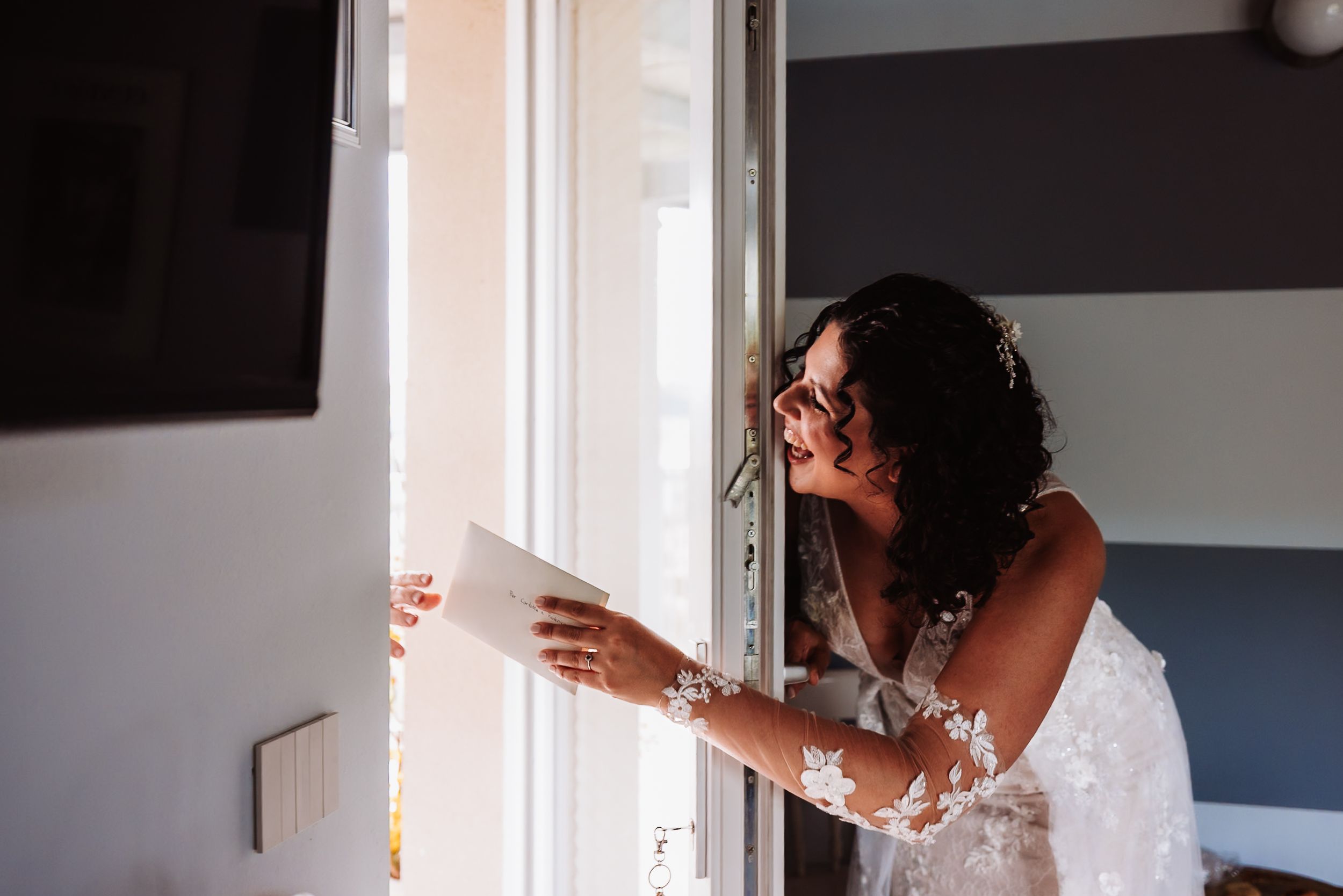 Bride in lace wedding dress and sunglasses reading a letter while leaning against an open door.