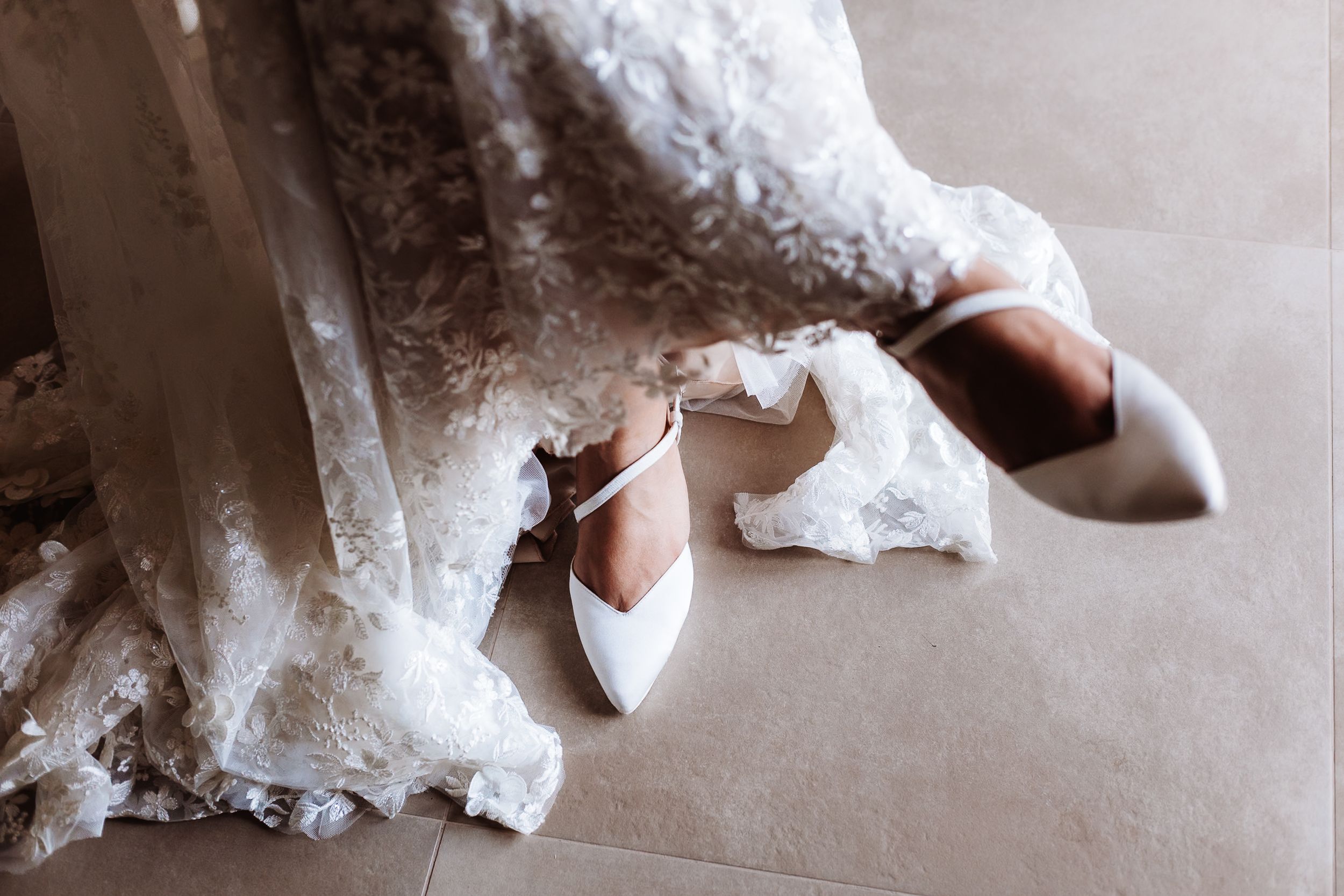White bridal heels with ankle strap placed on tile floor beside elegant lace wedding dress hem.