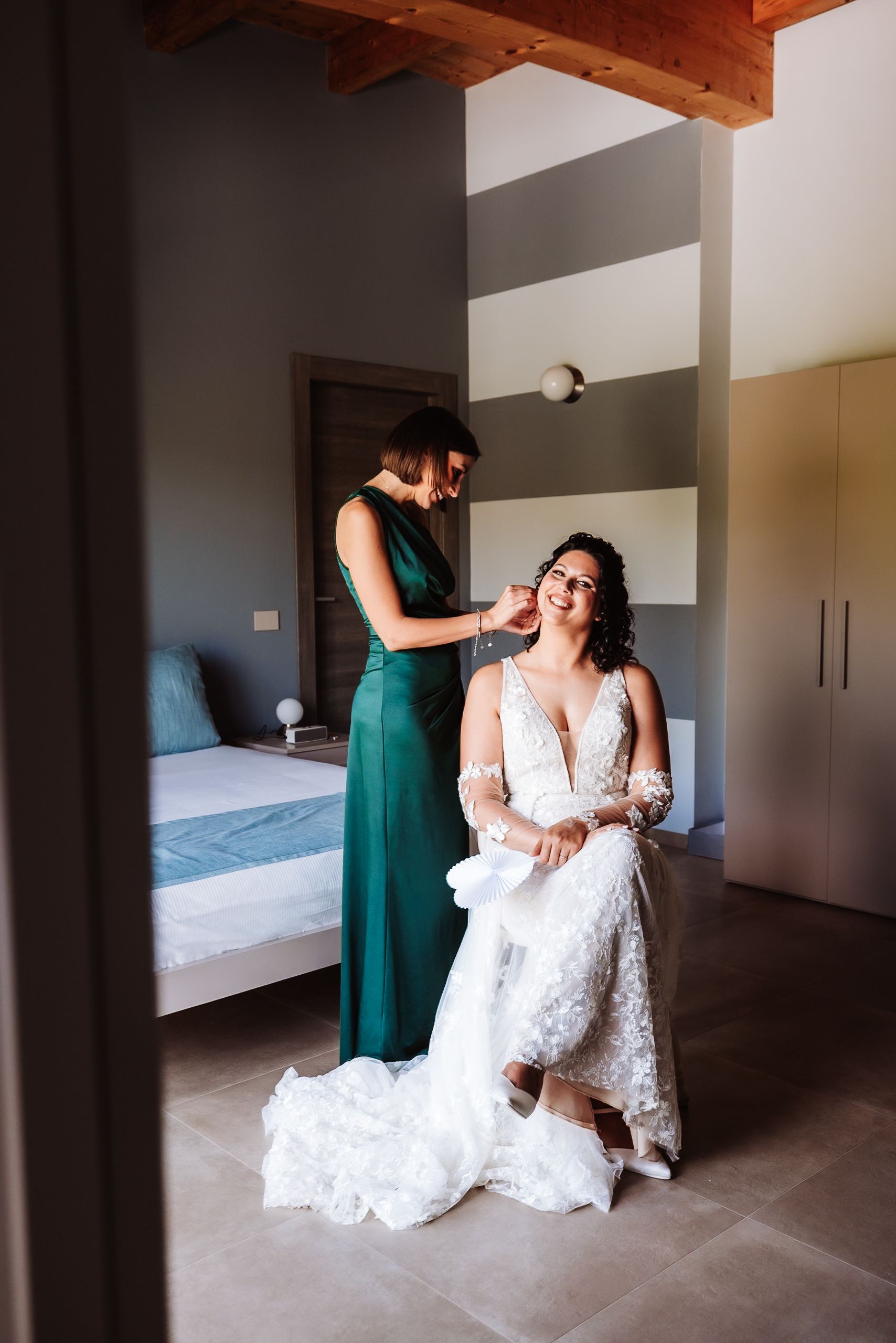 Bride in white lace gown seated while bridesmaid in green dress helps with final touches in elegant bedroom.