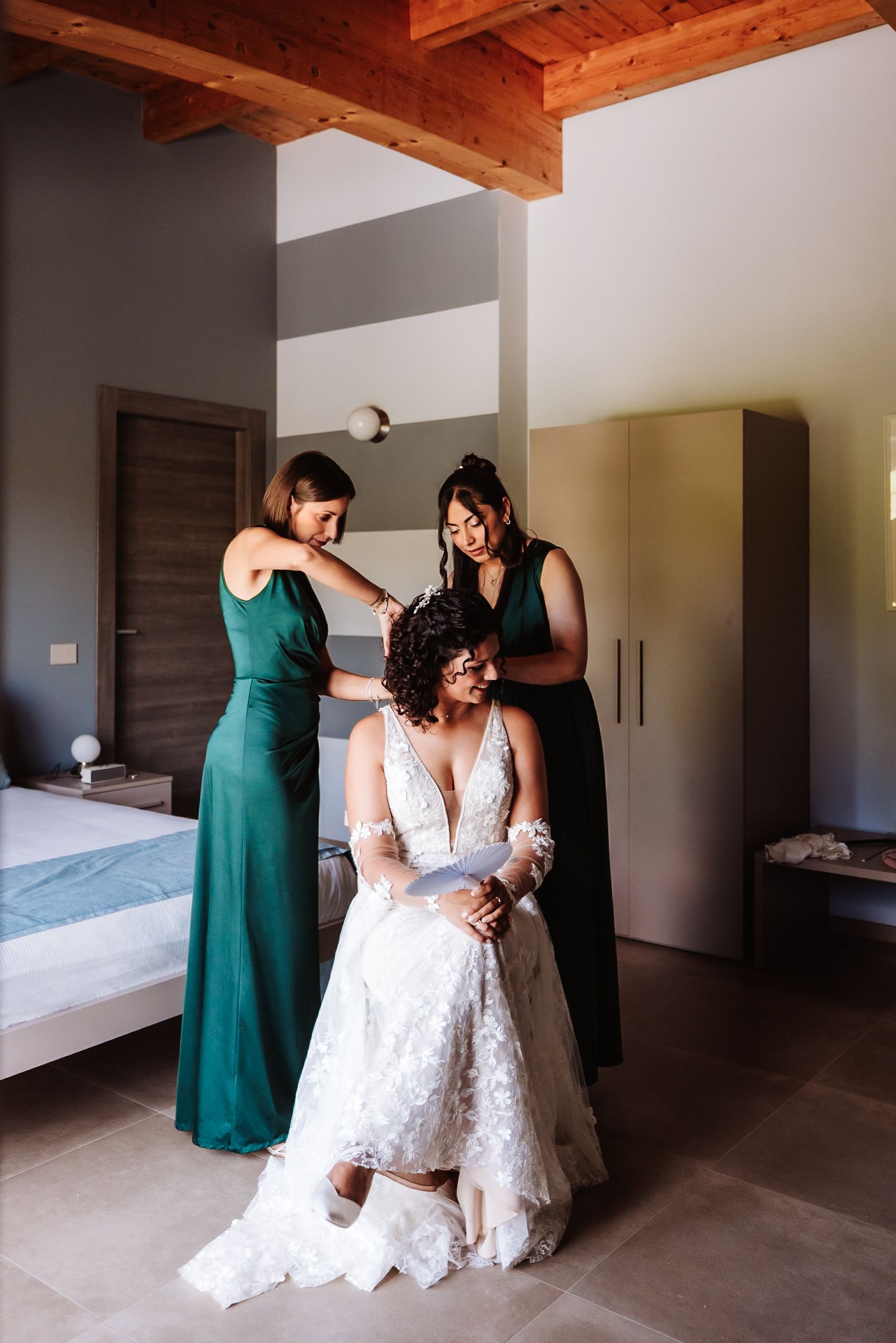 Bride in lace wedding gown seated while two bridesmaids in emerald green dresses help with hair in elegant bedroom.