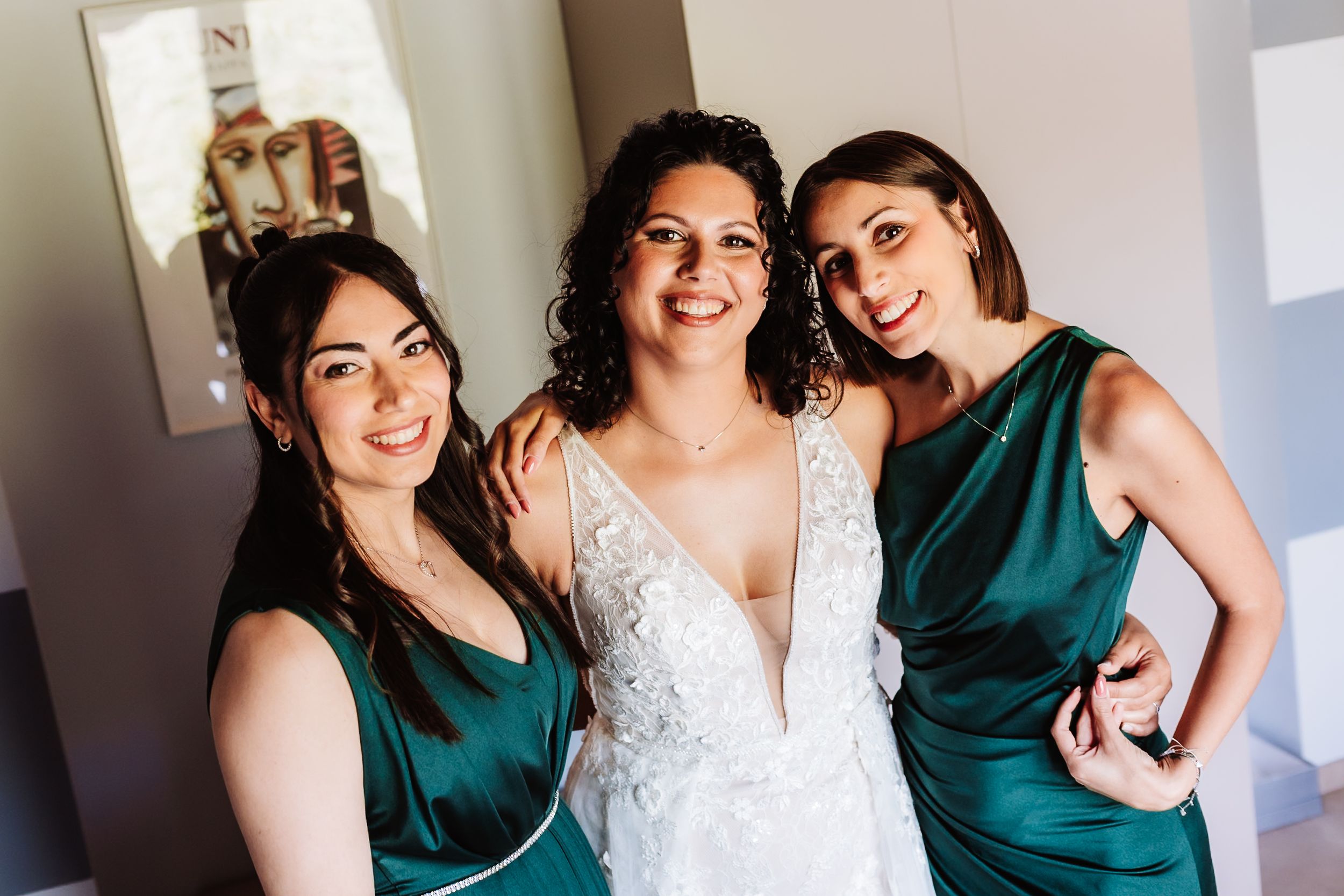 Bride in white lace gown poses with two bridesmaids in emerald green satin dresses at a wedding.
