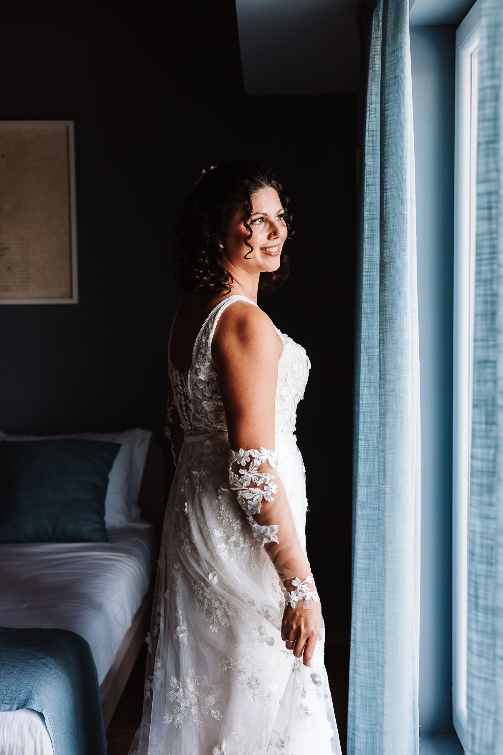 Bride in lace wedding gown smiling by window in dark elegant room, wearing floral sleeve detail.