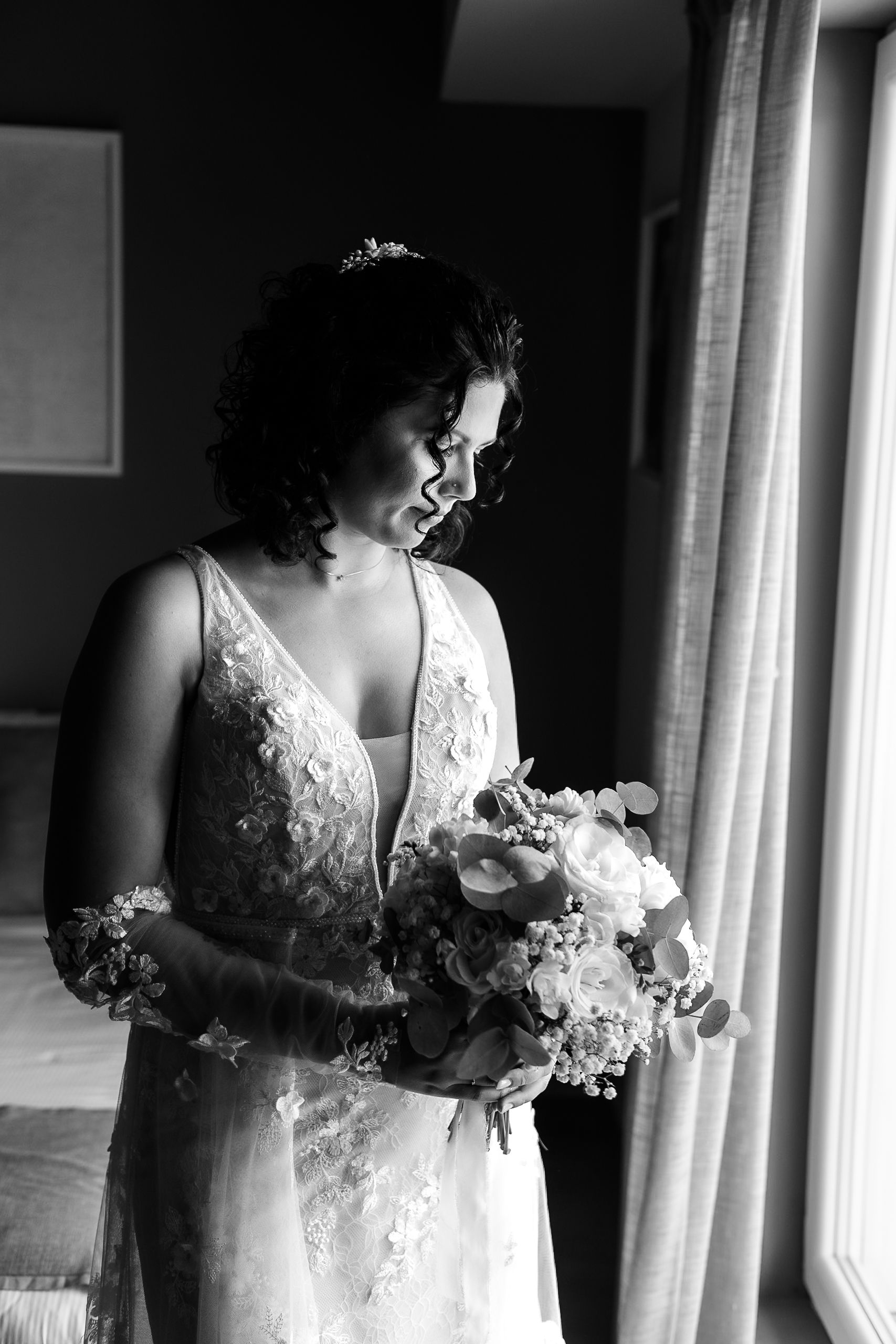 Black and white photo of a bride in lace gown holding a floral bouquet, standing by a window on her wedding day.