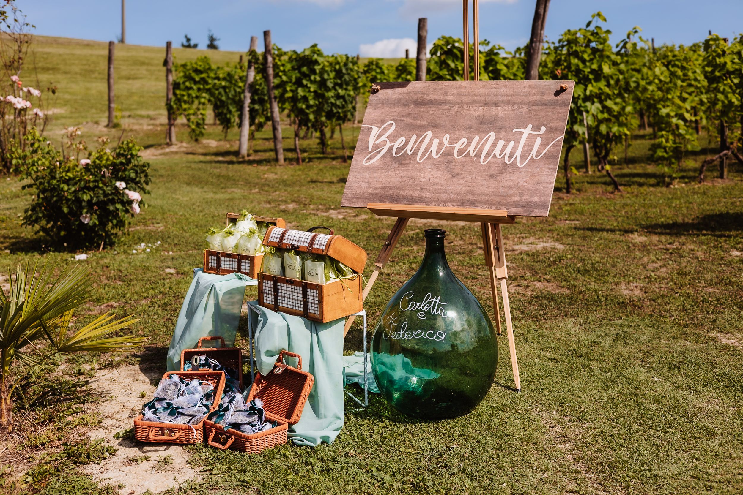 Rustic wedding welcome sign reading Benvenuti beside a large green glass demijohn and vintage suitcases in a vineyard.