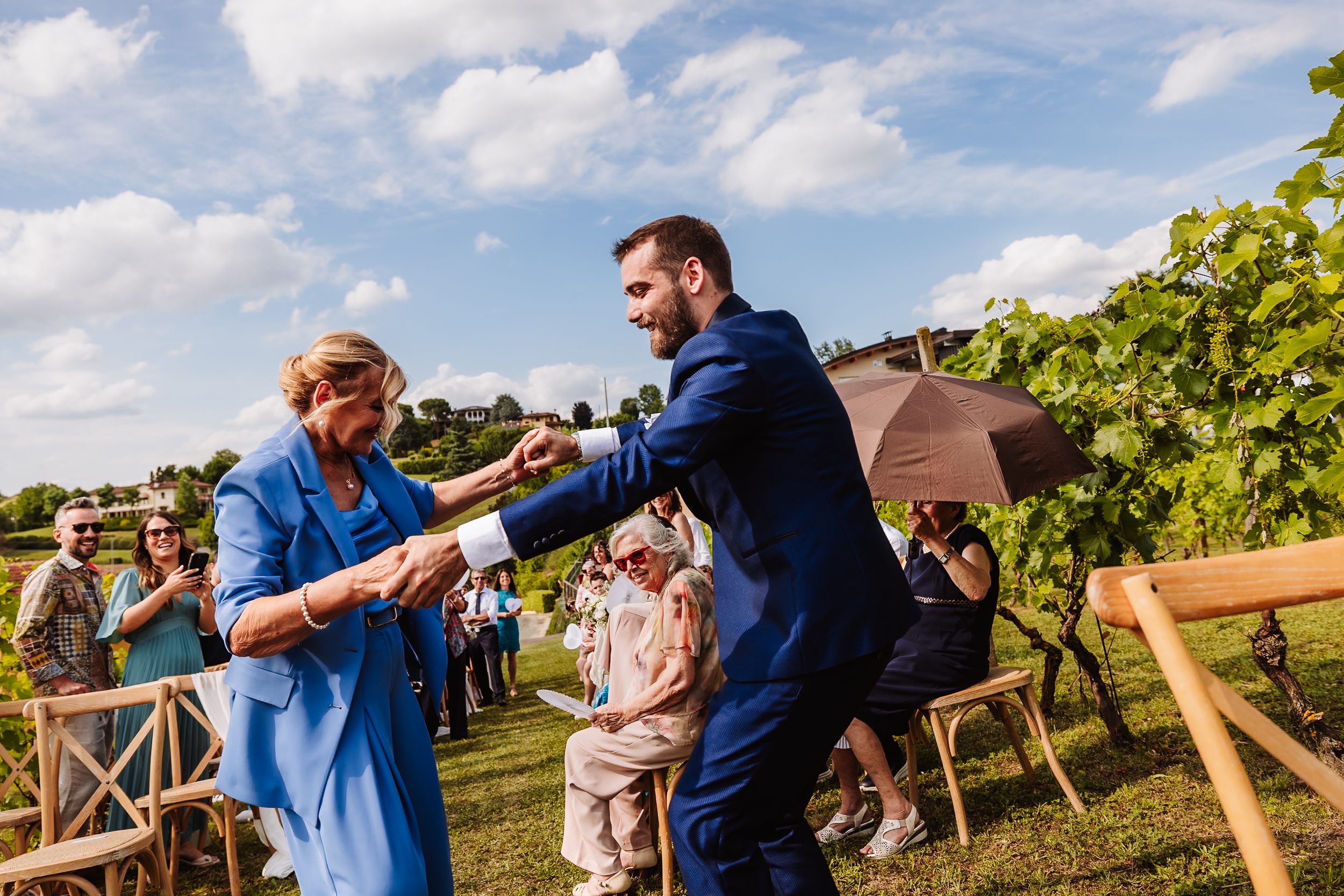 Groom in navy suit dancing with guest in blue dress at outdoor vineyard wedding ceremony.