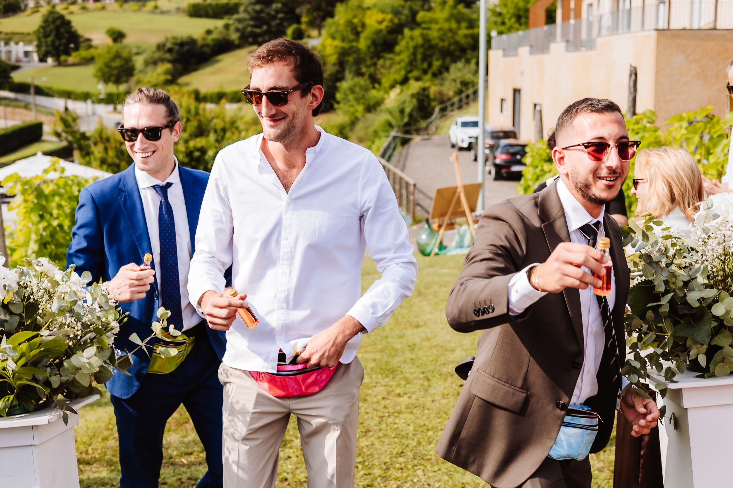 Men in sunglasses celebrating outdoors at a wedding, holding drinks and smiling on a sunny day.
