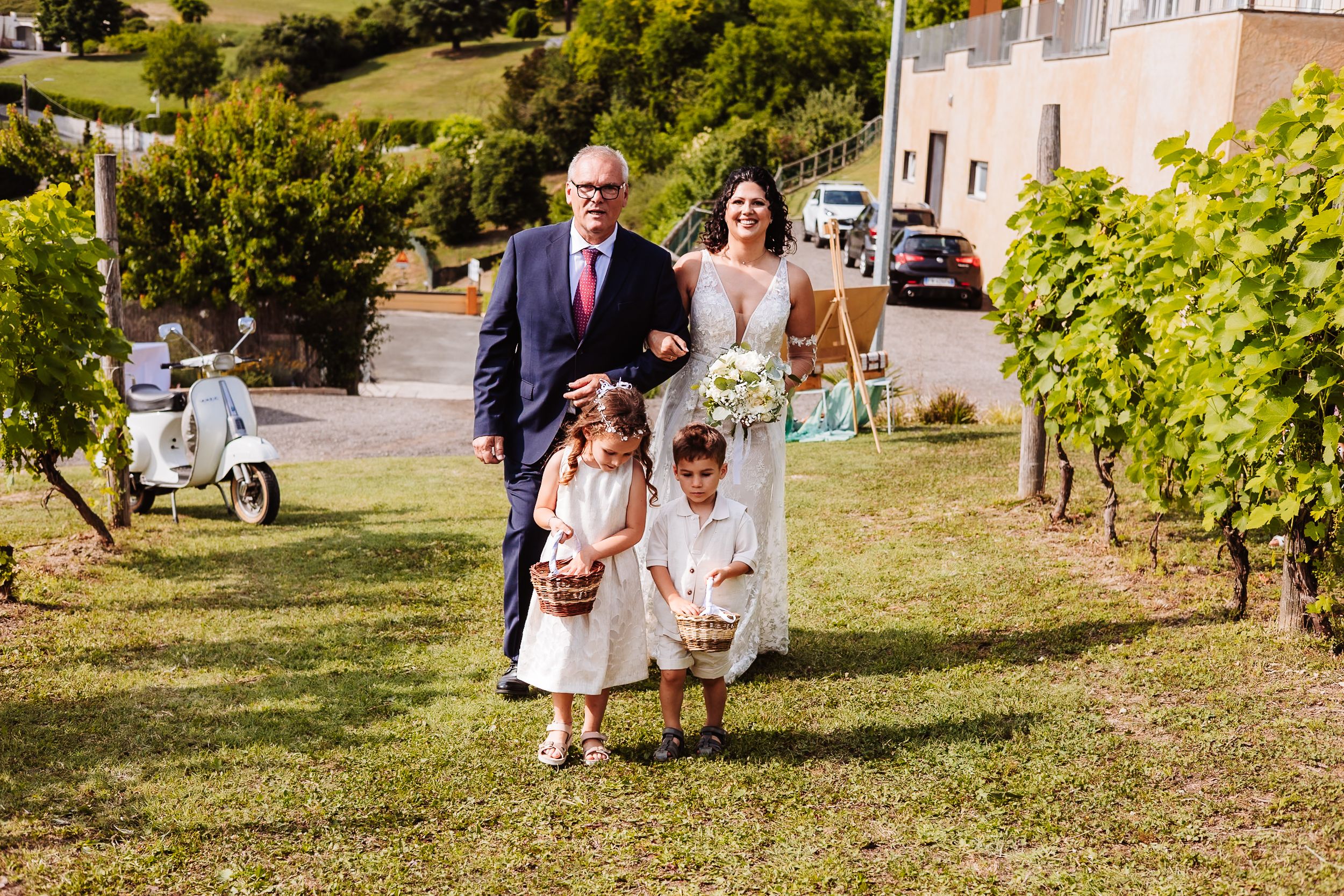 Bride walks with groom and two flower girls through vineyard at outdoor wedding ceremony on sunny day.