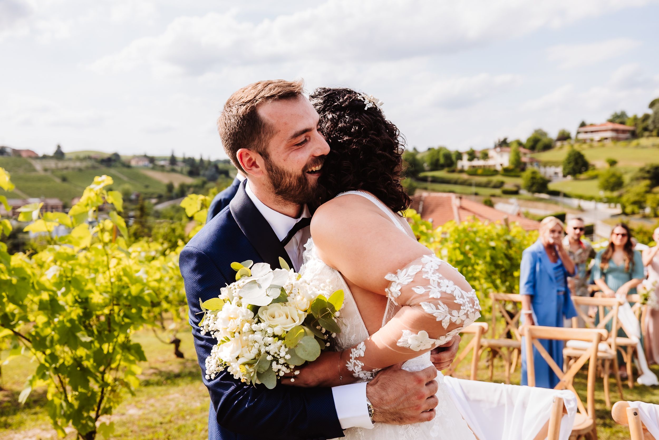 Newlywed couple sharing a romantic embrace at an outdoor vineyard wedding ceremony with guests in background.