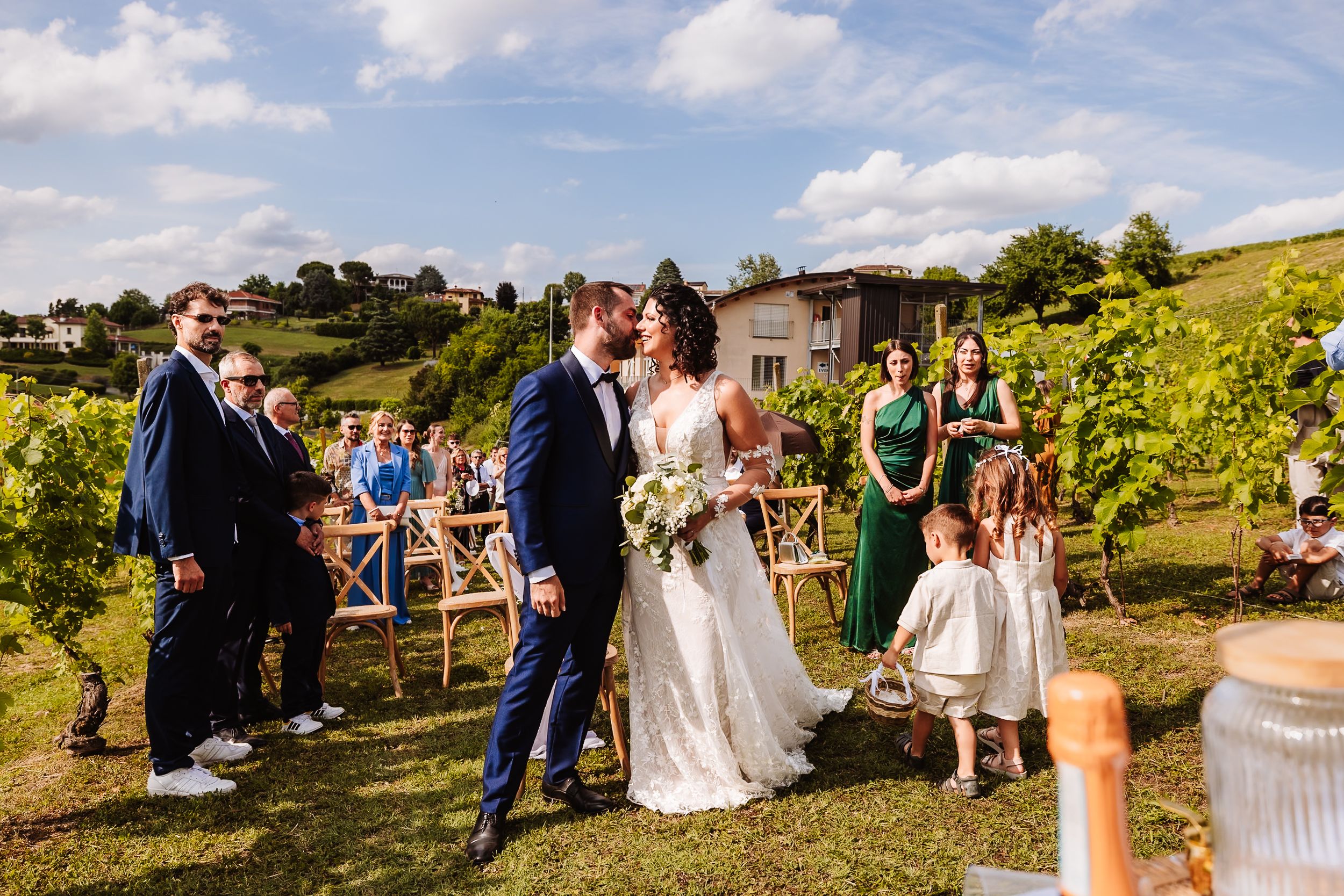 Bride and groom share a kiss at outdoor vineyard wedding ceremony with guests and flower girl nearby.
