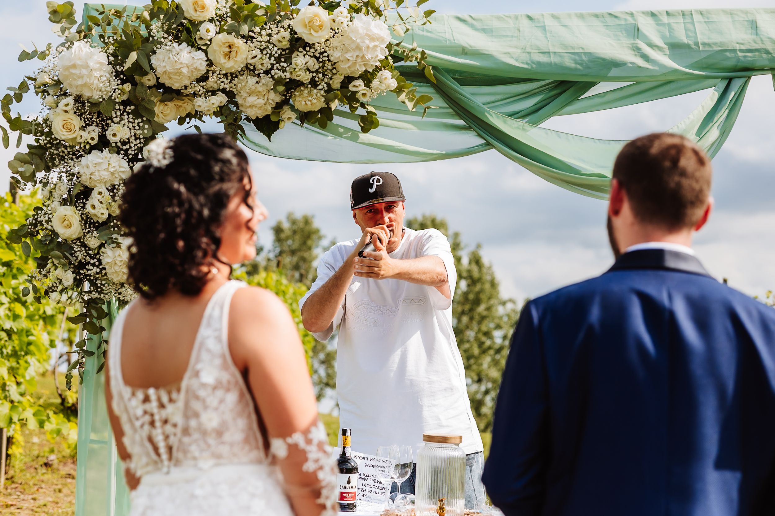 Officiant raises a toast under floral arch at outdoor vineyard wedding ceremony with bride and groom.