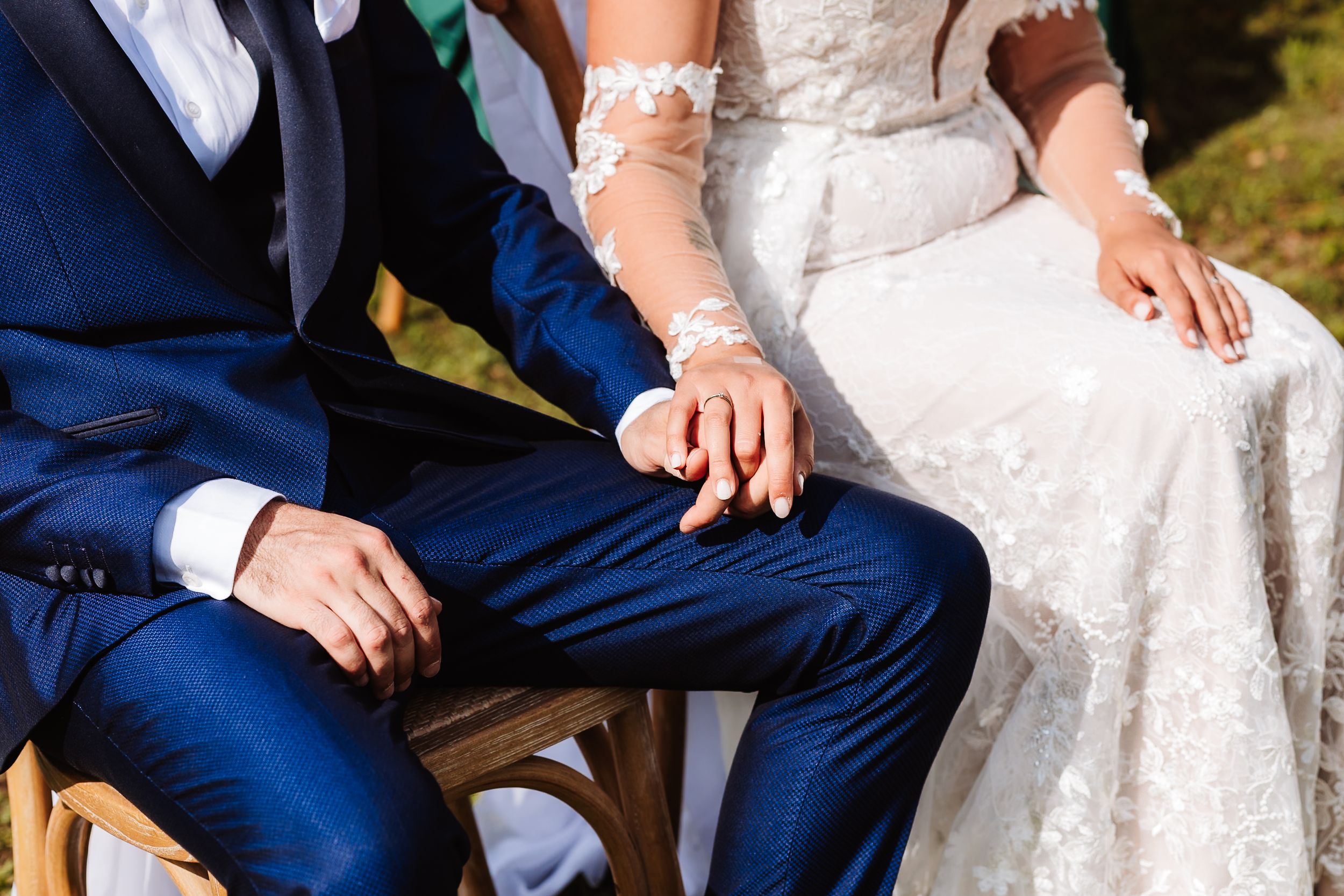Bride and groom holding hands at wedding ceremony, wearing elegant white lace dress and navy blue suit.