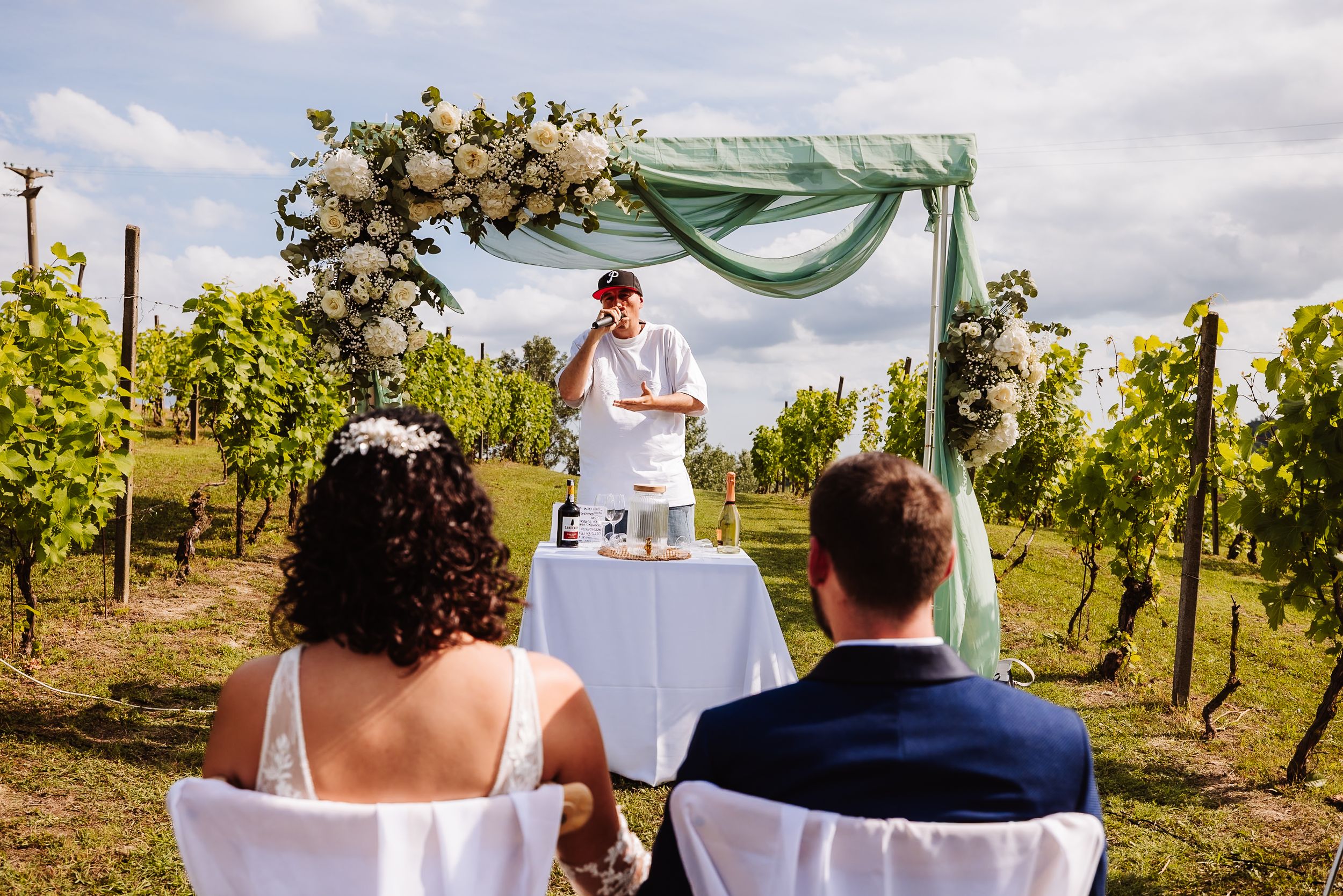 Outdoor vineyard wedding ceremony with couple seated before floral arch adorned with white roses and sage green draping.
