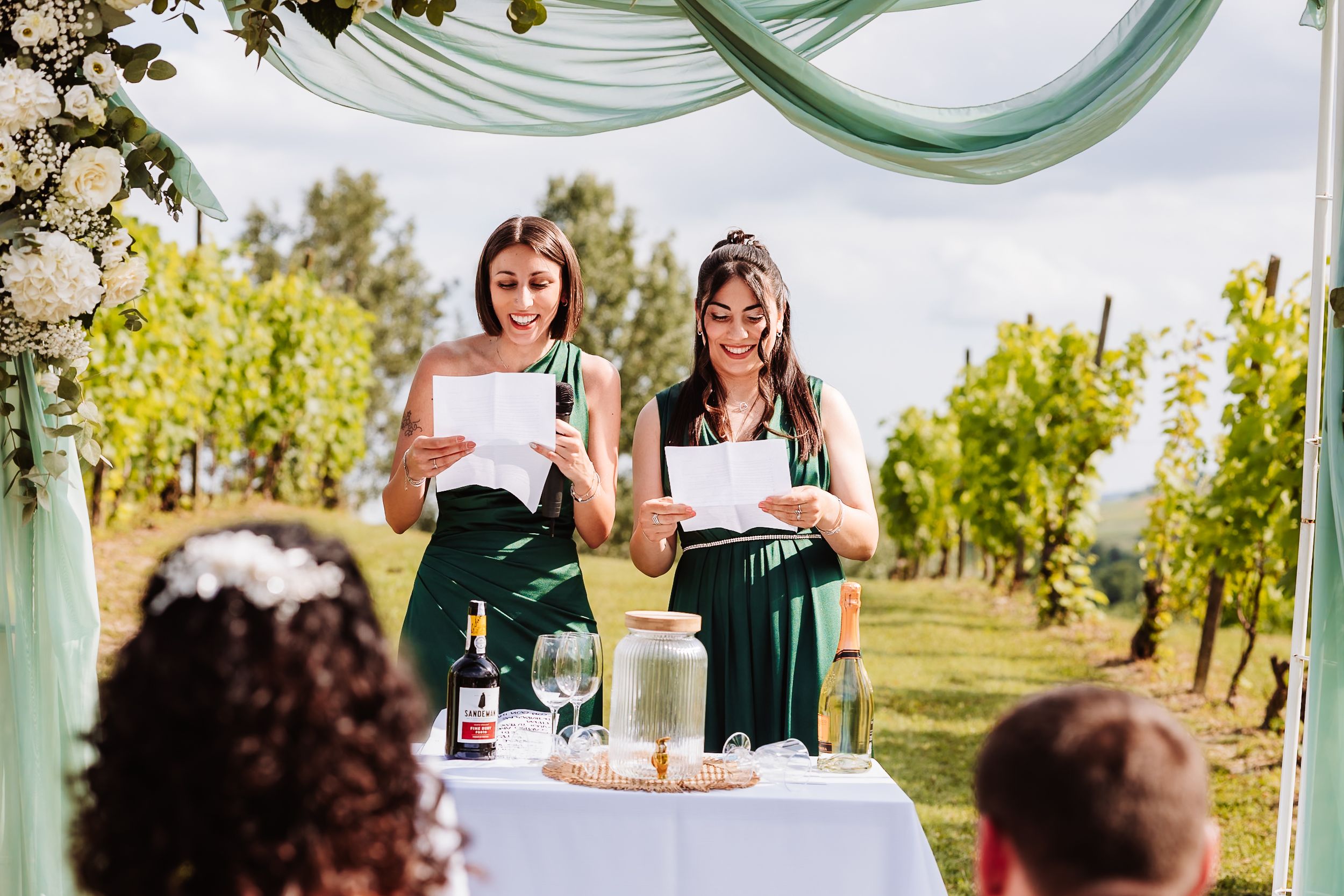 Two bridesmaids in green dresses reading wedding speeches at an outdoor vineyard ceremony under a floral arch.
