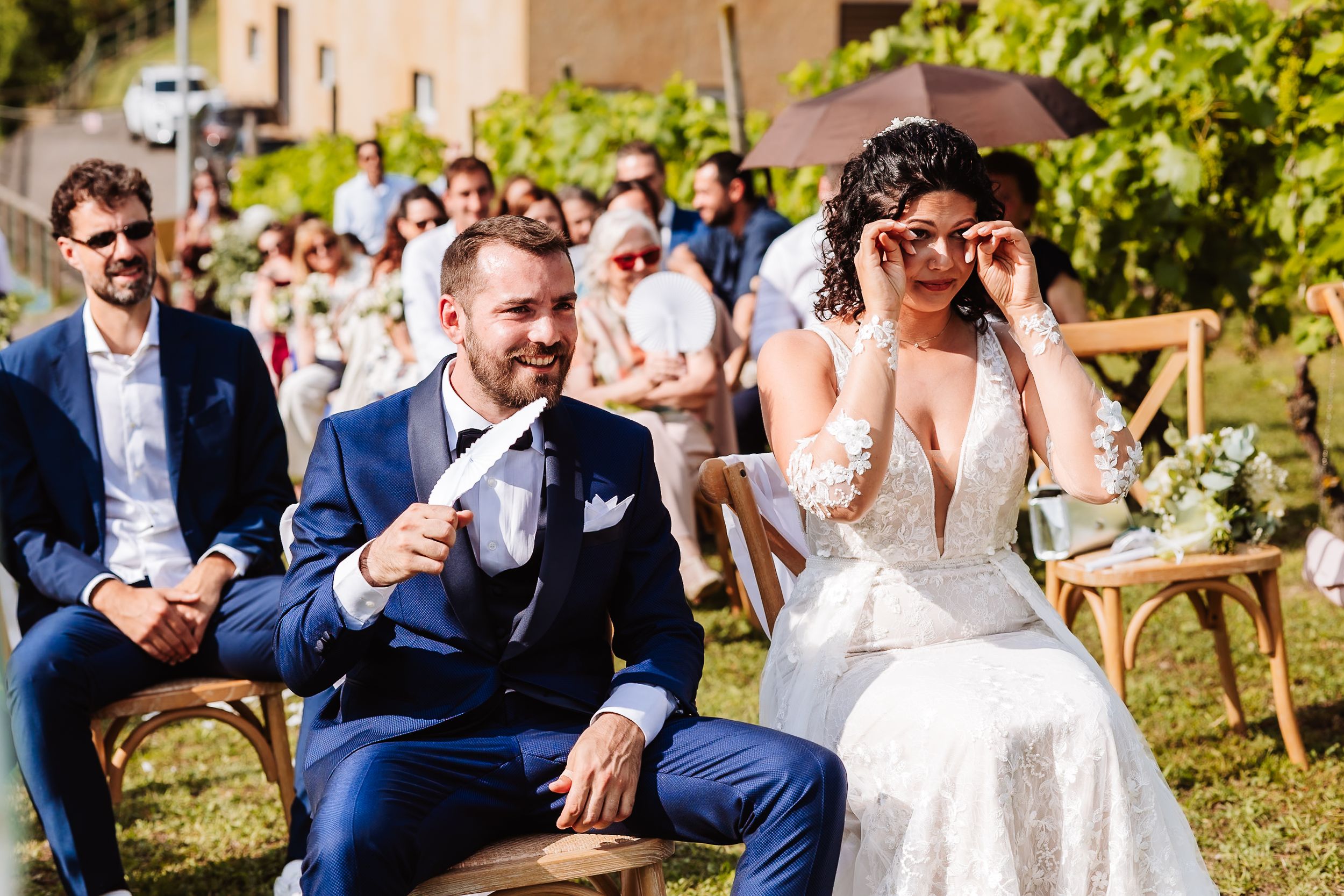 Bride and groom laughing during outdoor wedding ceremony, bride adjusting sunglasses, guests seated behind them.