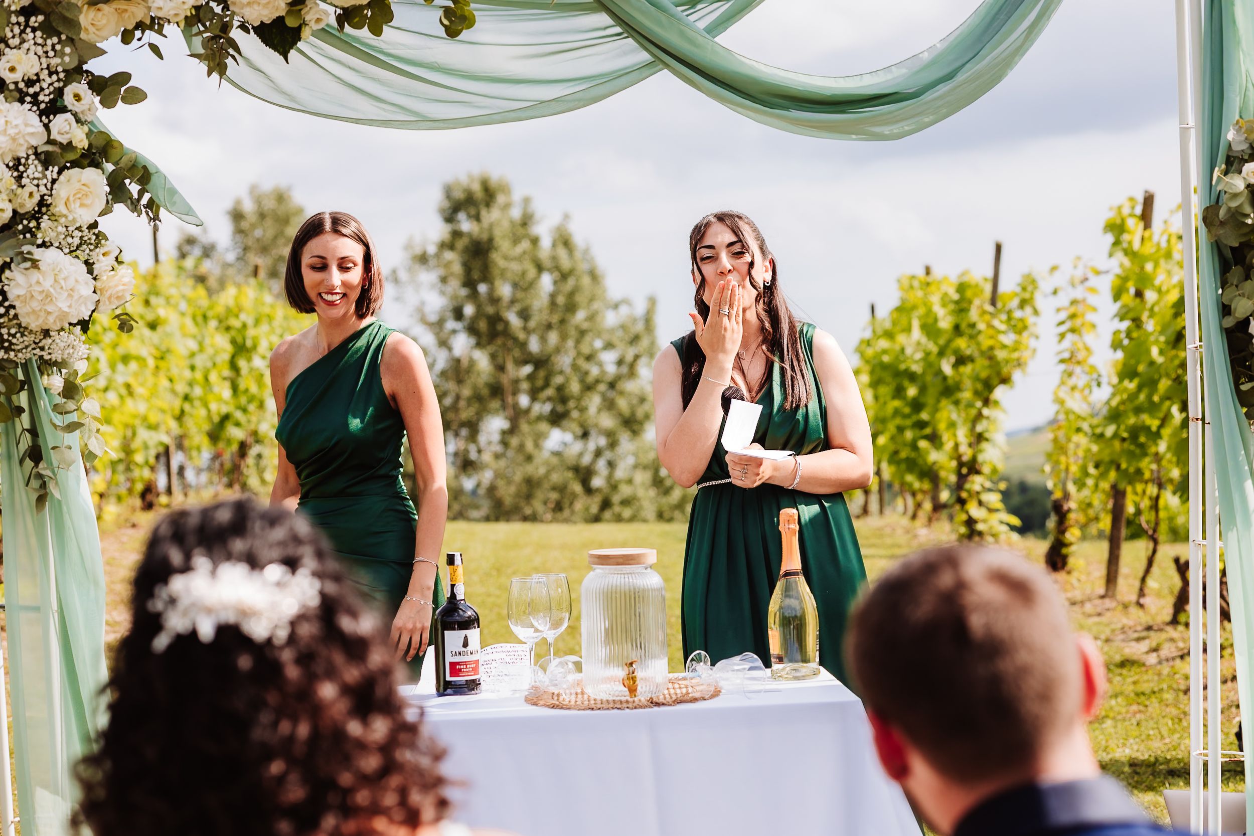 Two bridesmaids in green dresses give an emotional speech at an outdoor vineyard wedding ceremony.