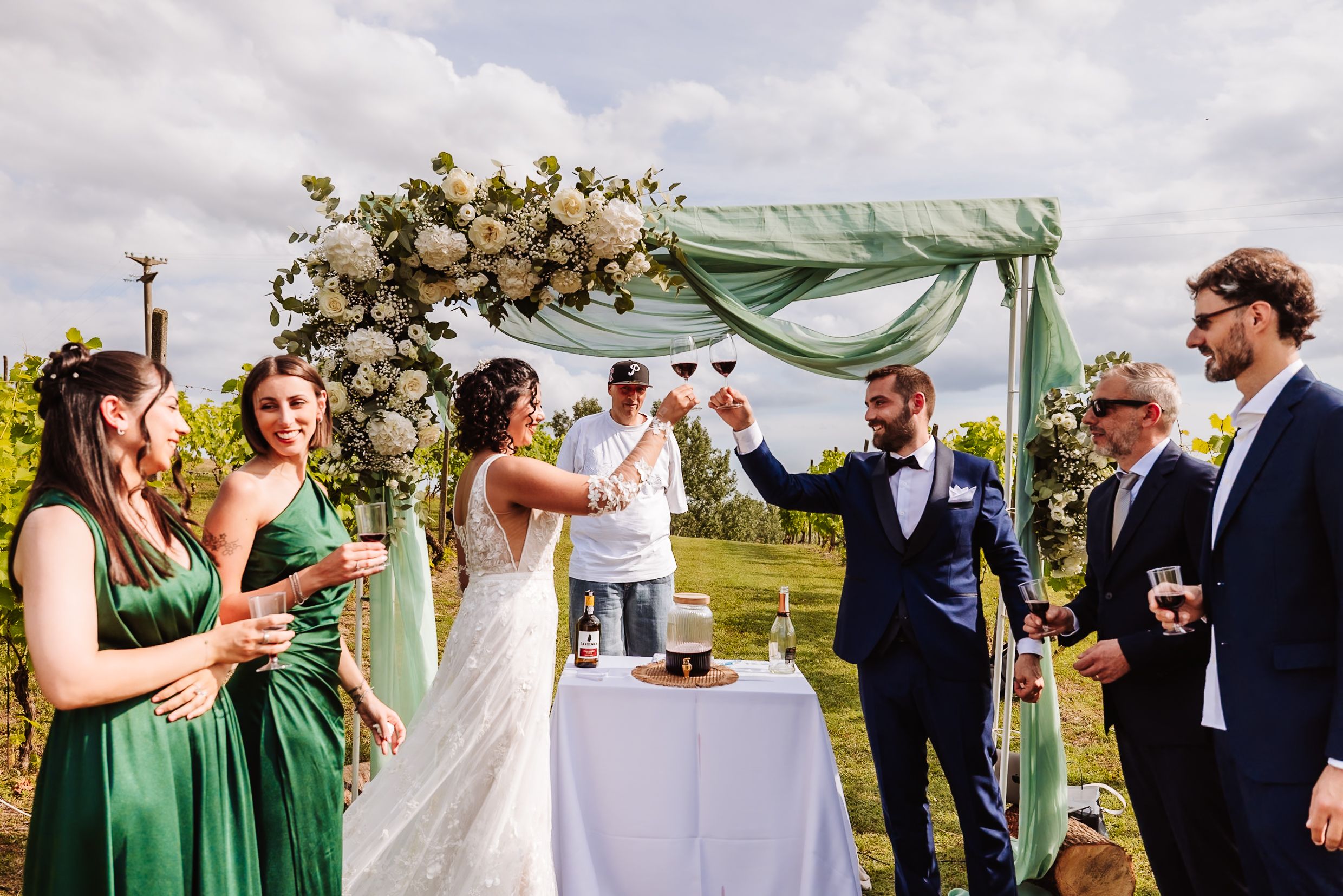 Outdoor wedding ceremony with bride and groom toasting under floral arch, flanked by guests in green dresses and suits.