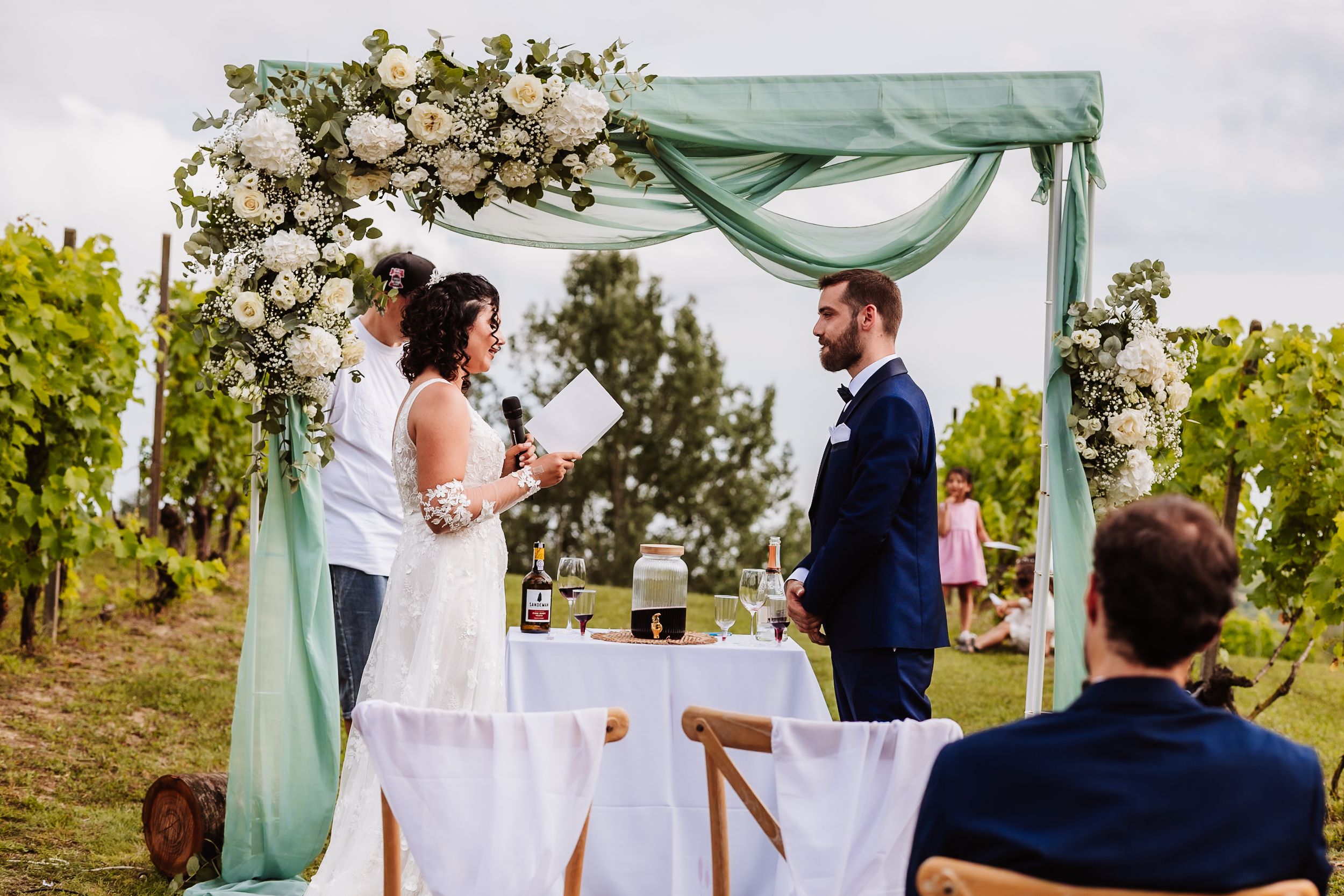 Outdoor vineyard wedding ceremony with couple at floral arch draped in sage green fabric, surrounded by lush greenery.