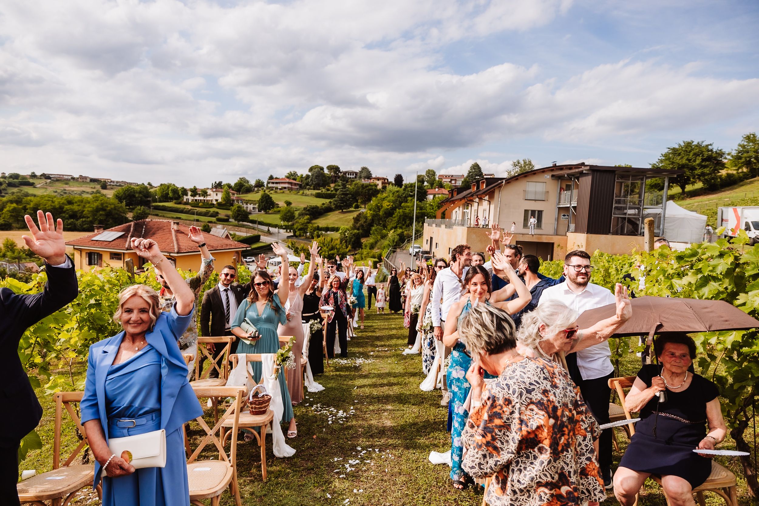 Outdoor wedding ceremony in a vineyard with guests waving, surrounded by lush greenery and rolling hills.