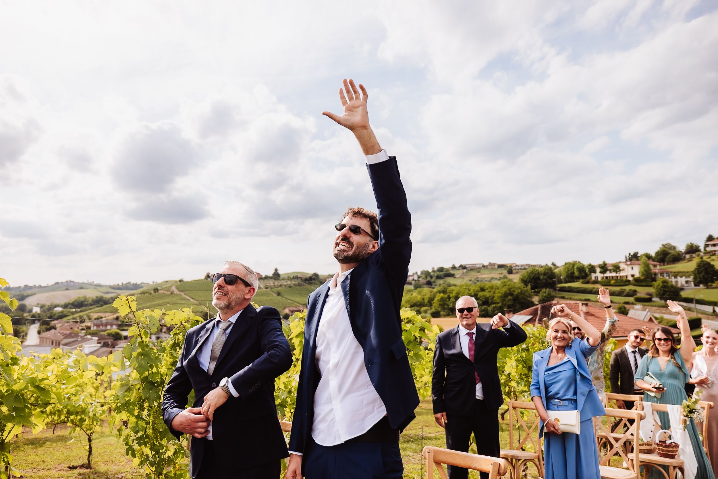 Man in navy suit raising hand at outdoor vineyard wedding ceremony with guests in background.