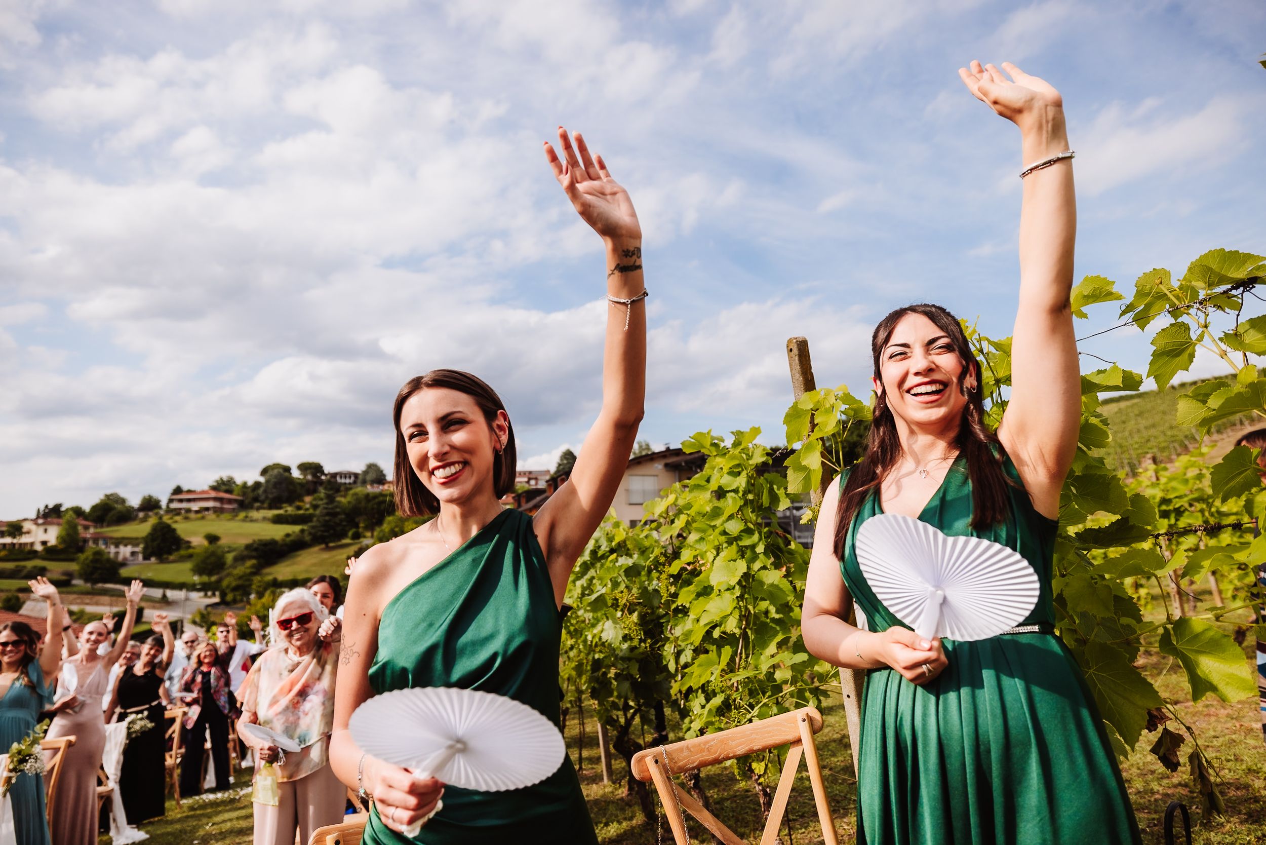 Two bridesmaids in green dresses celebrate joyfully at an outdoor vineyard wedding ceremony on a sunny day.