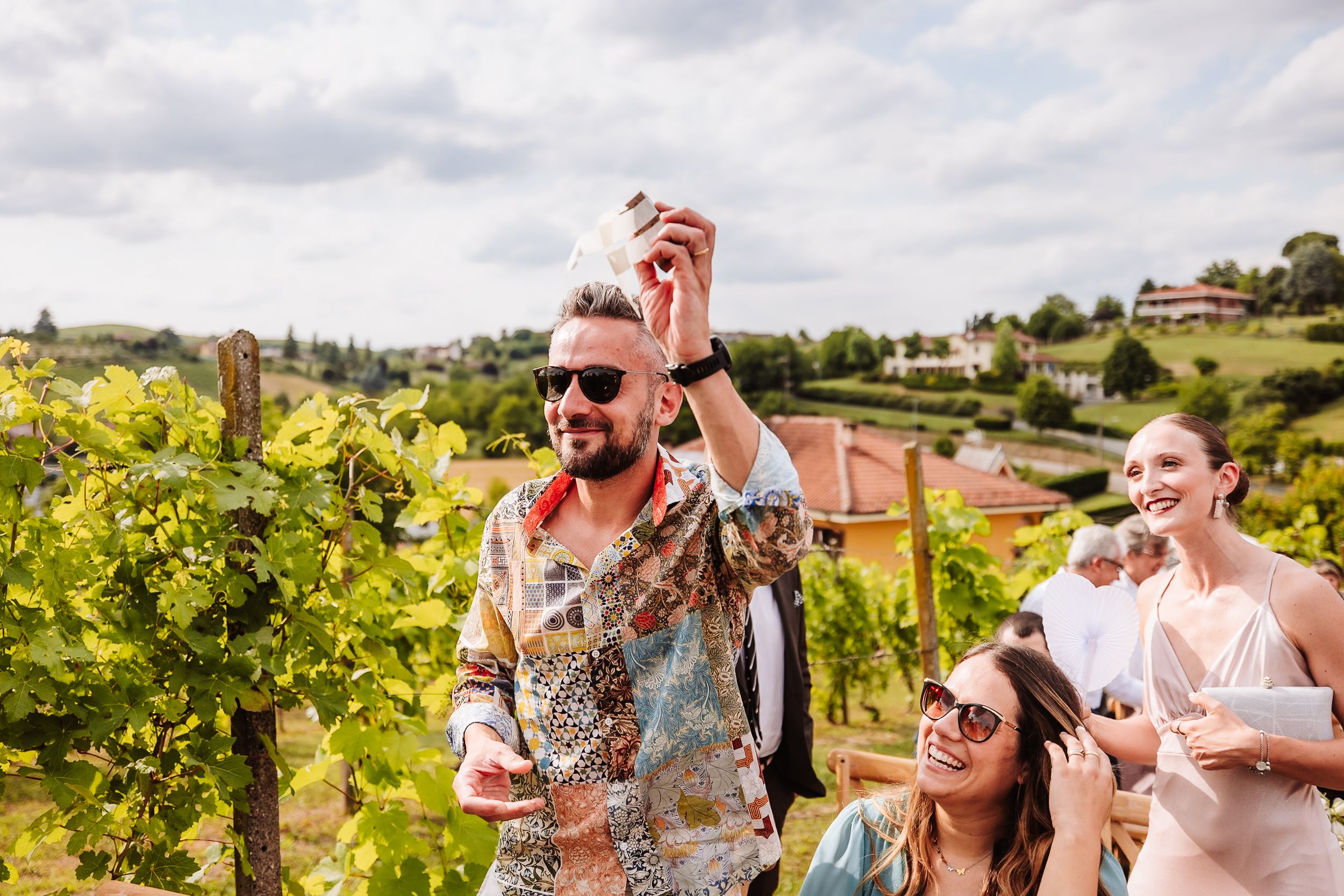 People celebrating in a vineyard, man in floral shirt raising a glass, rolling green hills in the background.