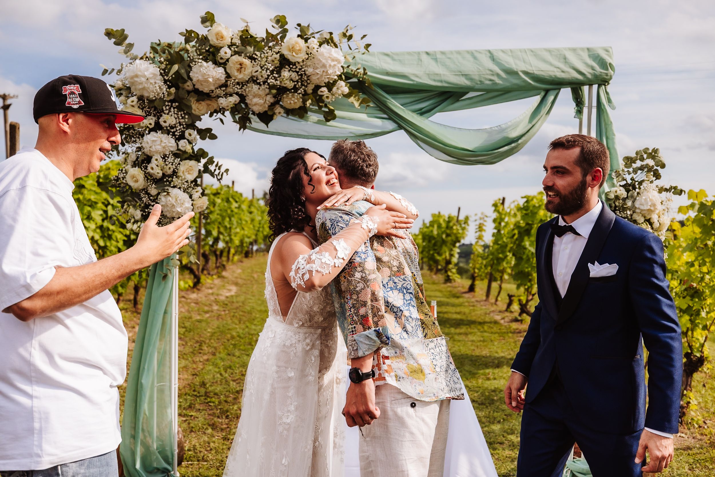 Bride and groom share a hug with a guest under a floral arch at an outdoor vineyard wedding ceremony.