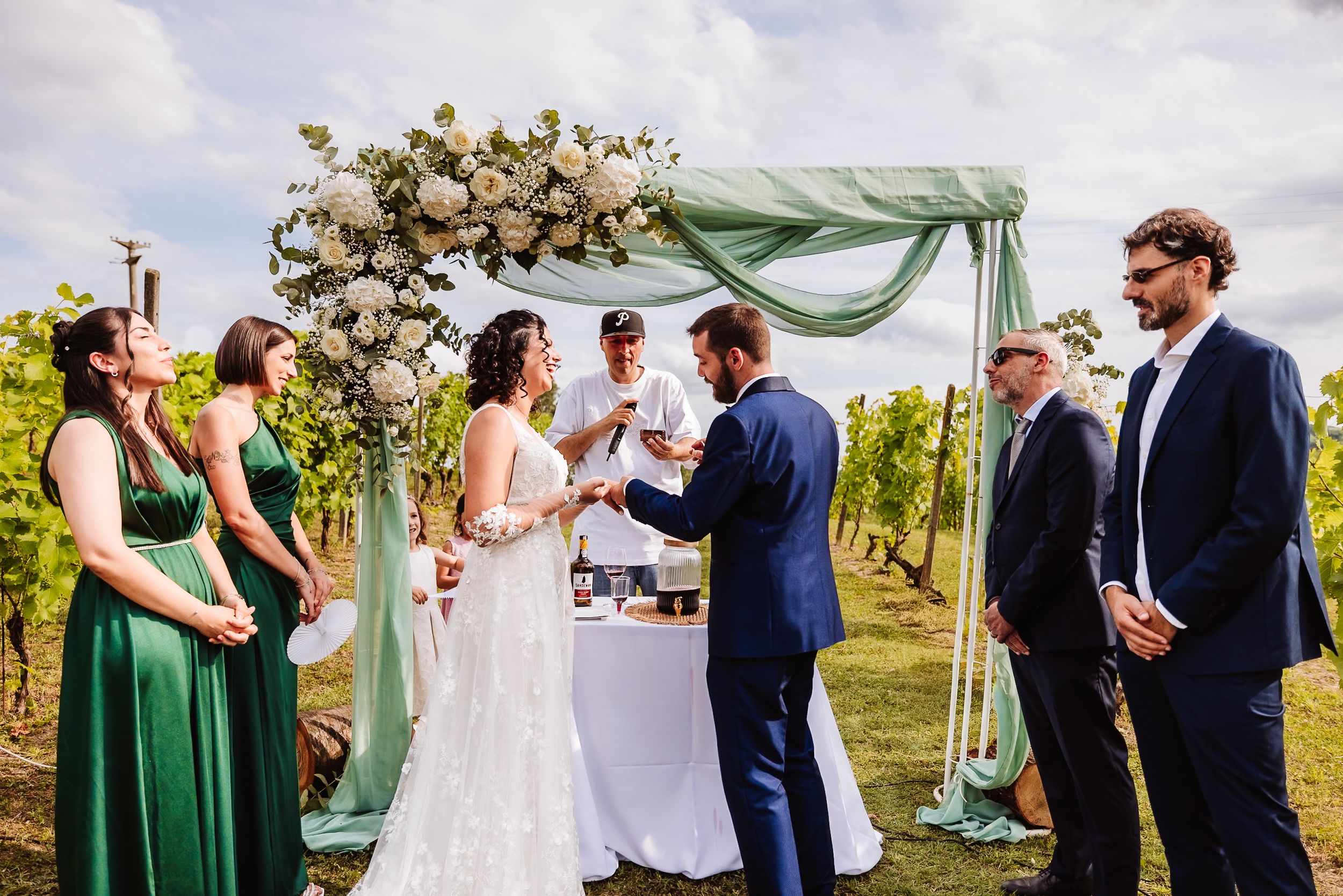 Outdoor vineyard wedding ceremony under green draped arch with white floral arrangements, bride, groom, and wedding party.