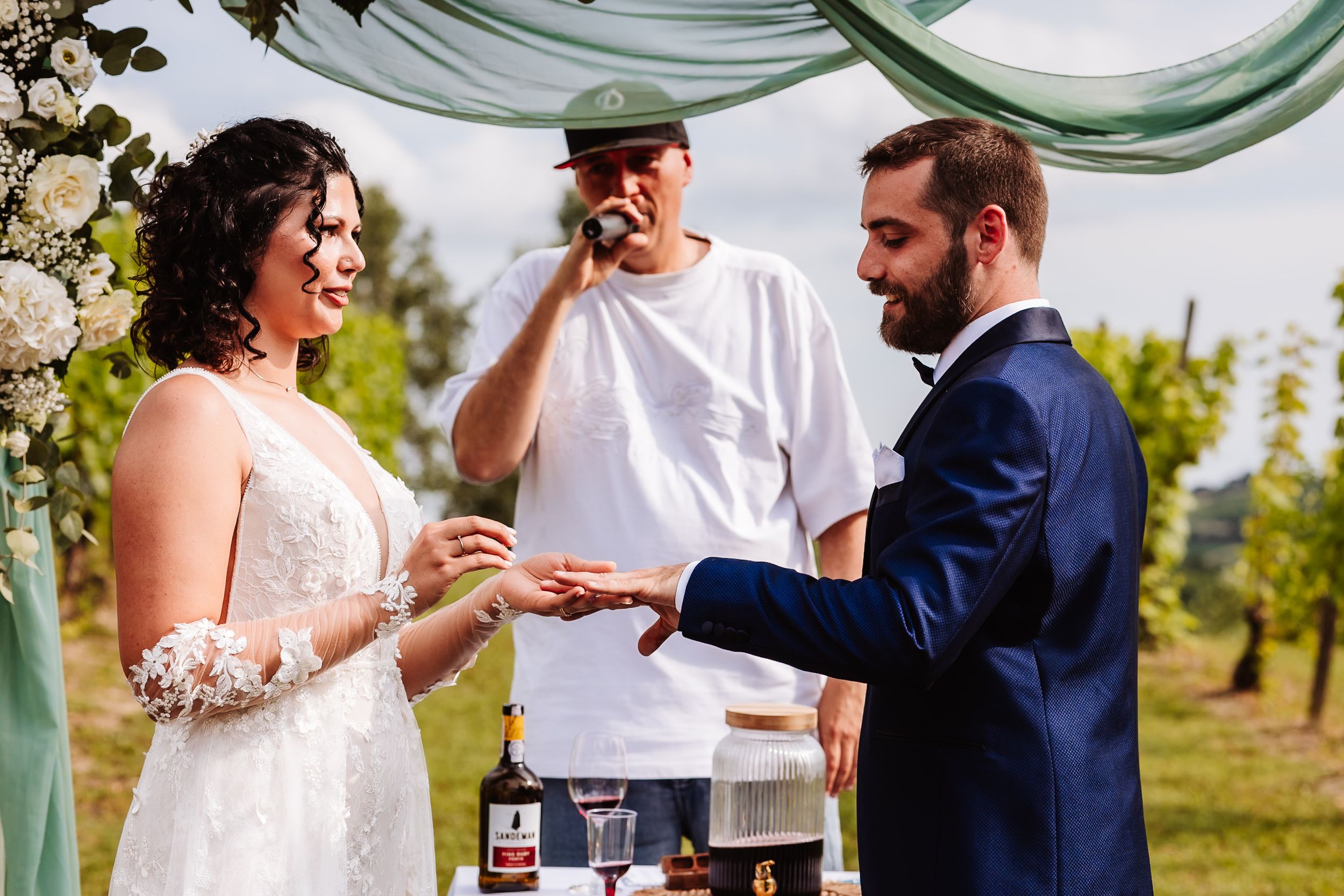 Bride and groom exchange rings outdoors at a vineyard wedding ceremony with an officiant in a white shirt.
