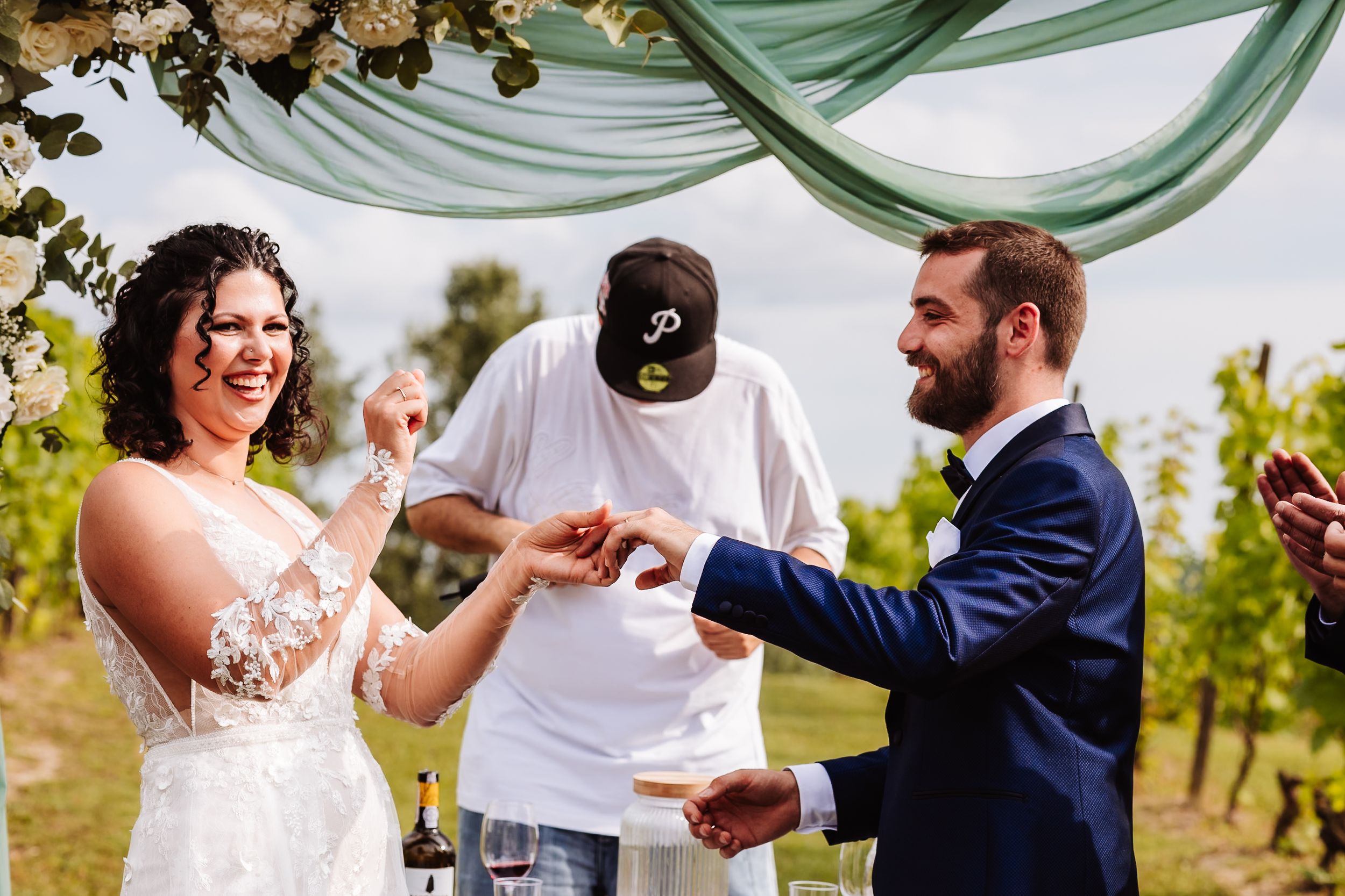 Bride and groom exchanging rings during outdoor vineyard wedding ceremony with officiant under floral arch.