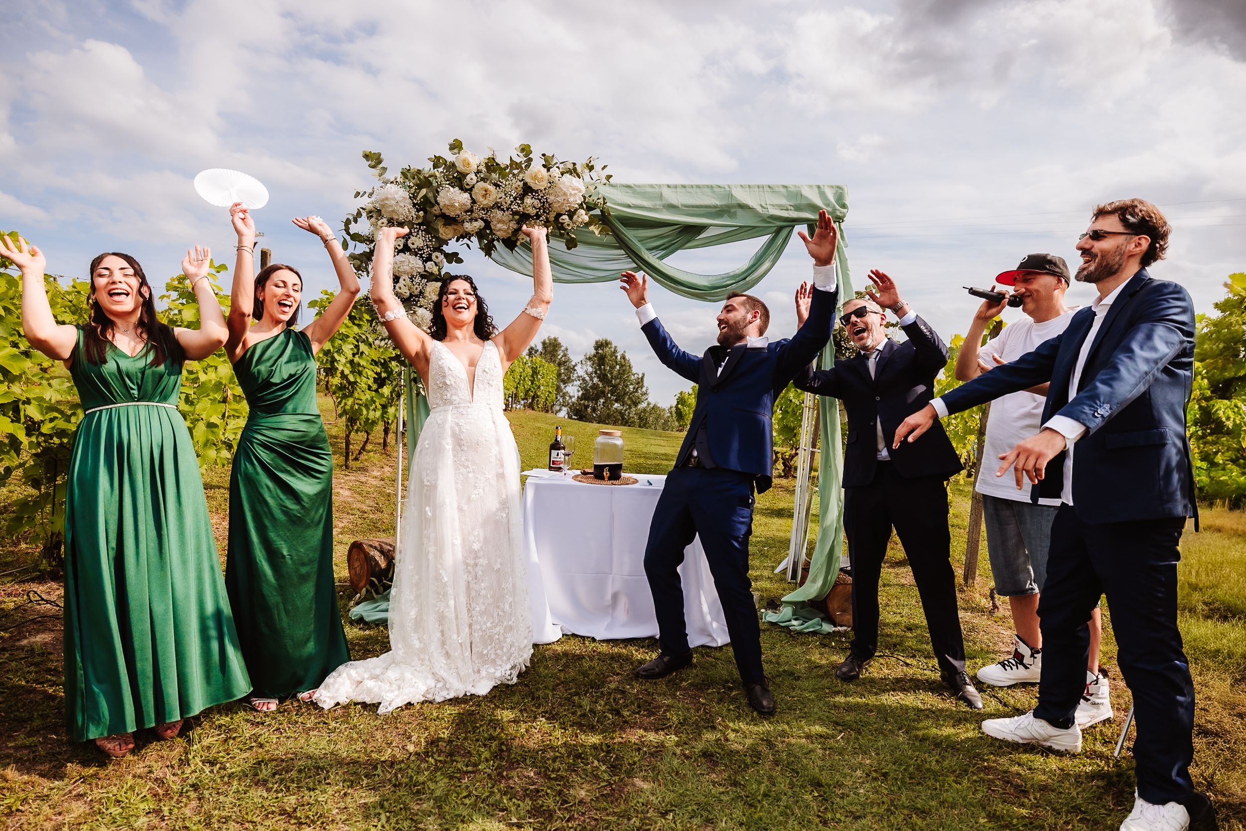 Joyful wedding celebration outdoors in vineyard with bride, groom, green-dressed bridesmaids cheering under floral arch.