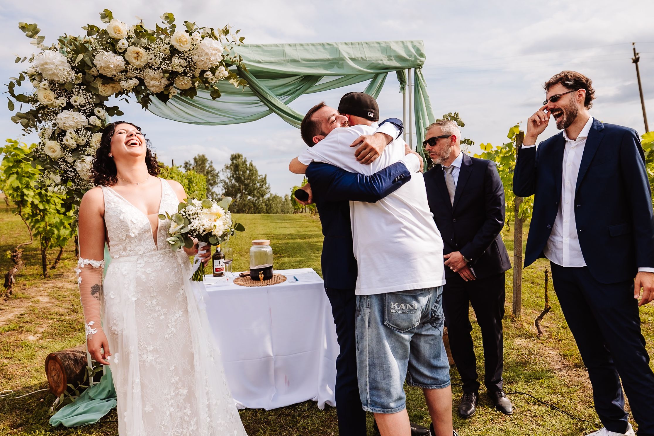 Joyful wedding ceremony in vineyard with bride holding bouquet, groom hugging guest under floral arch.