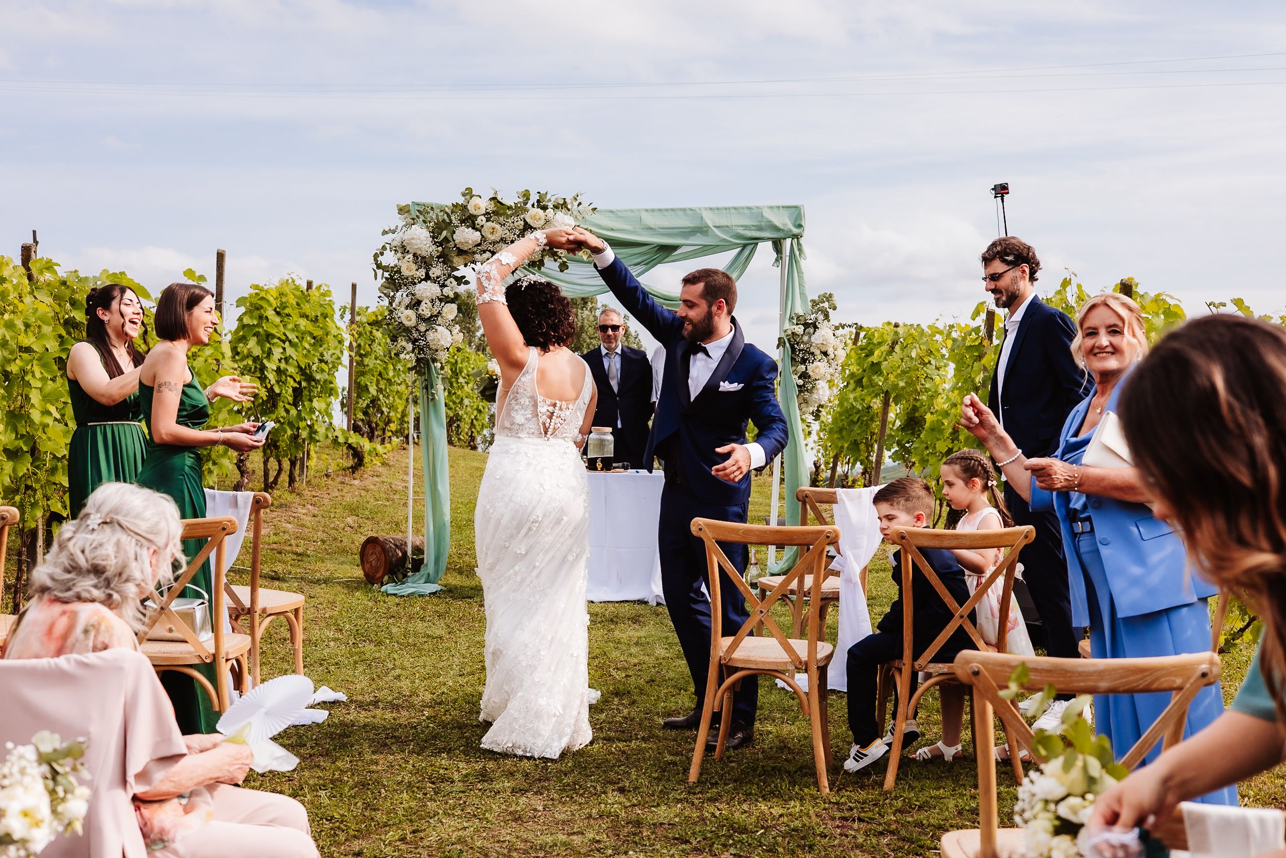 Joyful newlyweds raise hands at outdoor vineyard wedding ceremony with guests applauding in elegant summer setting.