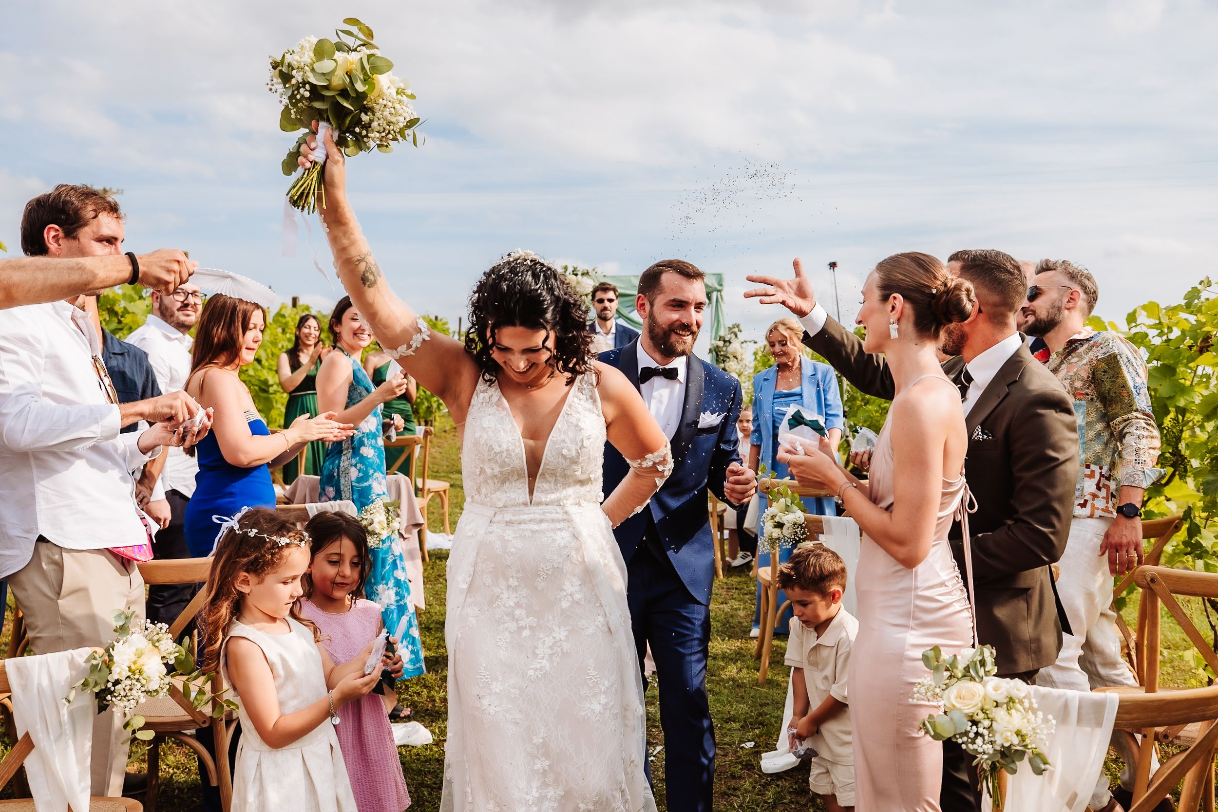Joyful newlyweds walk down the aisle as guests celebrate at an outdoor vineyard wedding ceremony.