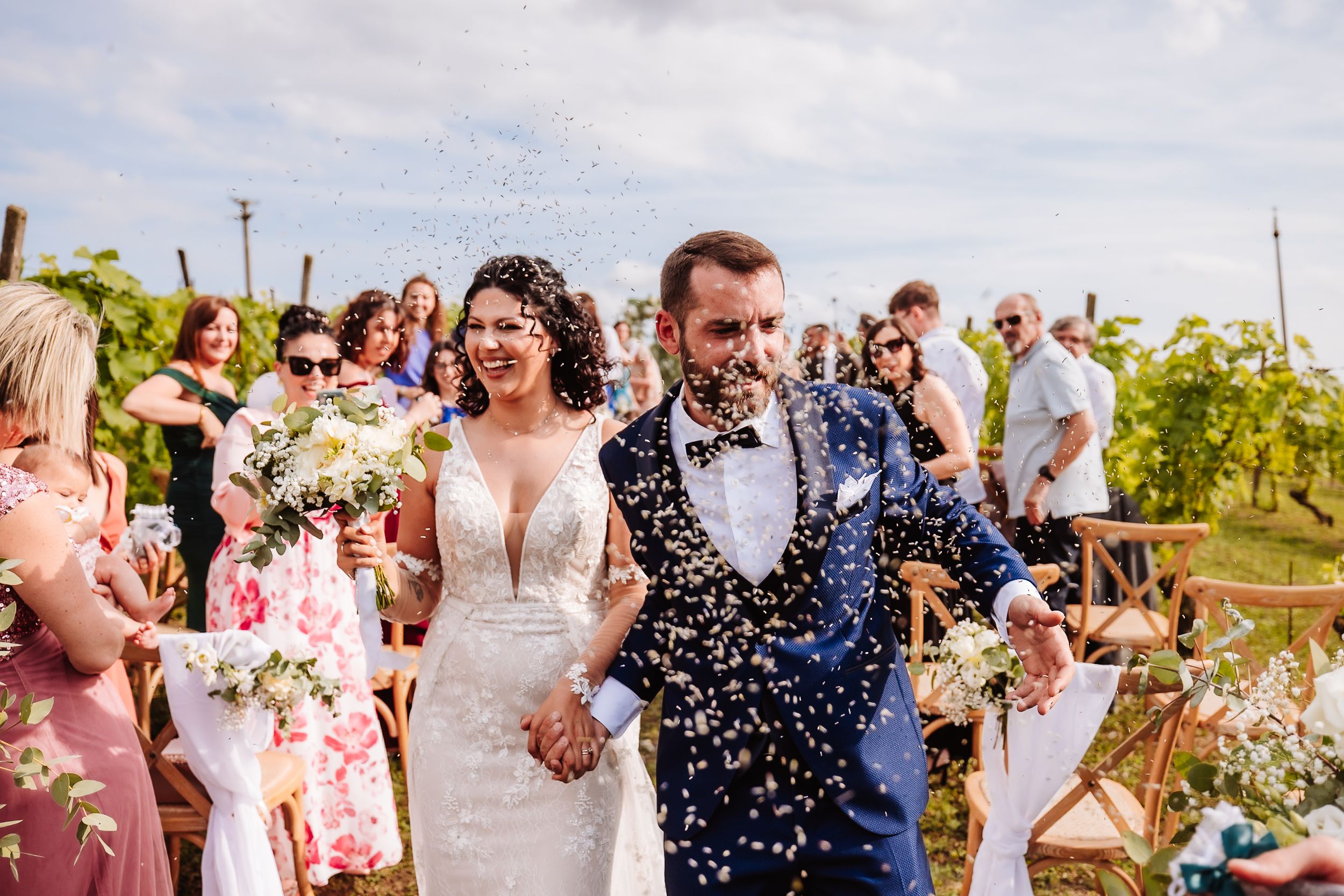Bride and groom walk down vineyard aisle as guests toss white confetti during outdoor wedding ceremony.